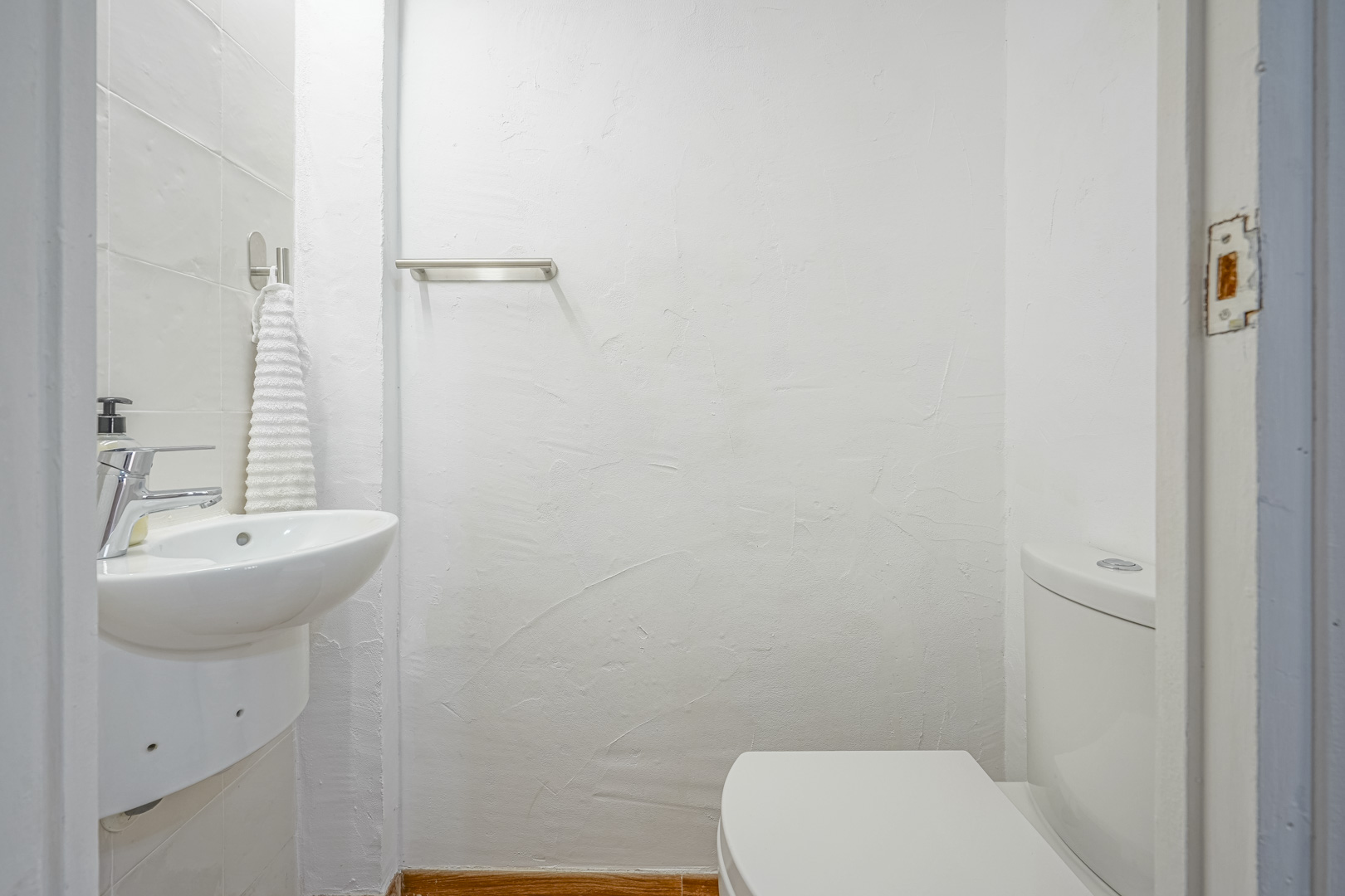 Compact guest toilet featuring textured white walls, a wall-mounted ceramic sink with chrome fixtures, and a stainless steel towel rail.