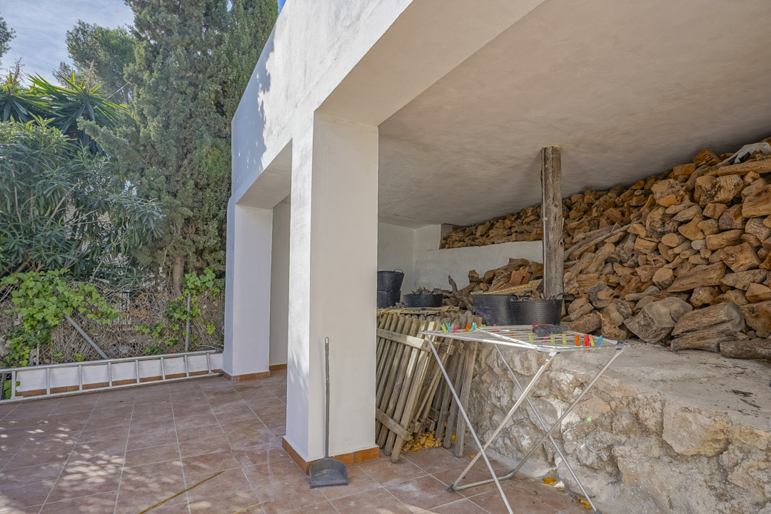 Covered outdoor storage area featuring a tiled terrace, white structural pillars, and a large woodpile stacked against a natural stone wall.
