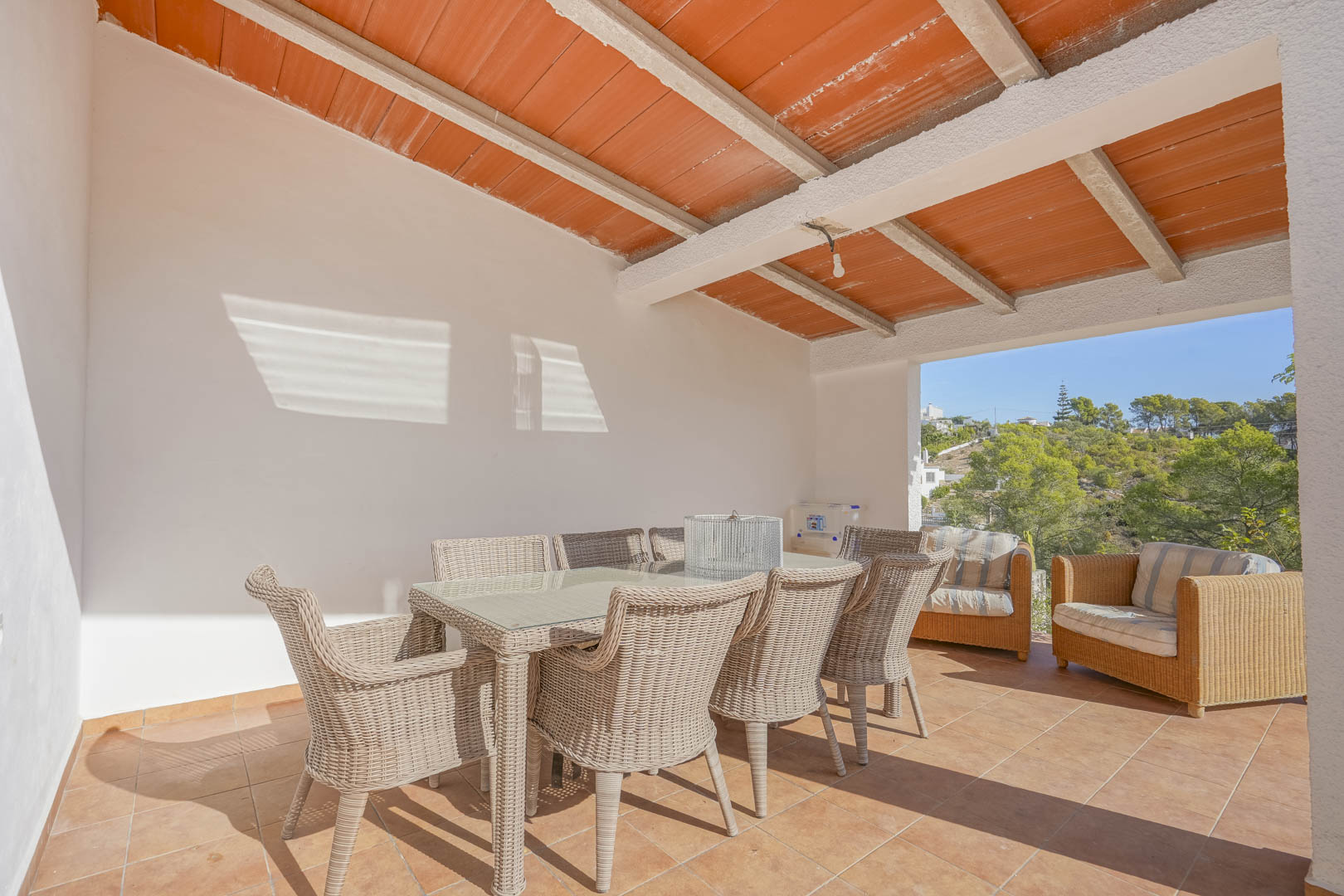 Covered terrace featuring a beamed ceiling with terracotta tiles, a wicker dining set for six, and open views of the surrounding pine-covered hills.