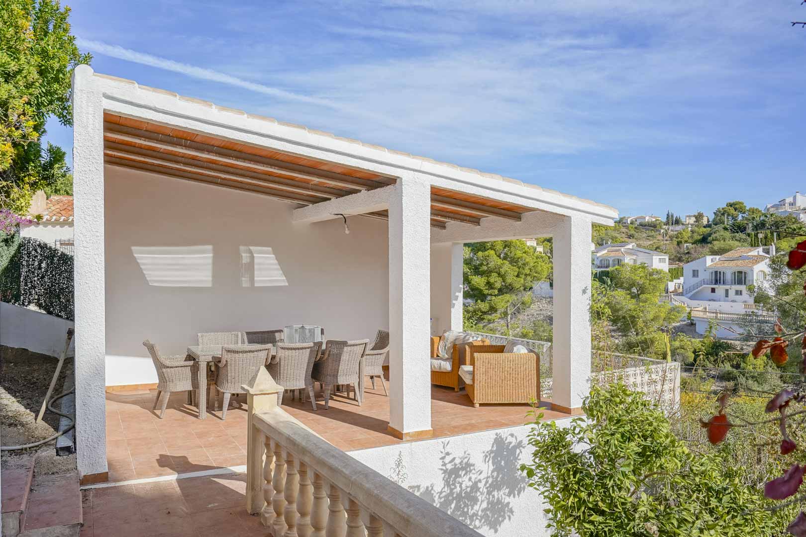 Elevated covered terrace featuring a beamed ceiling, wicker dining and lounge sets, and views over the Mediterranean landscape.