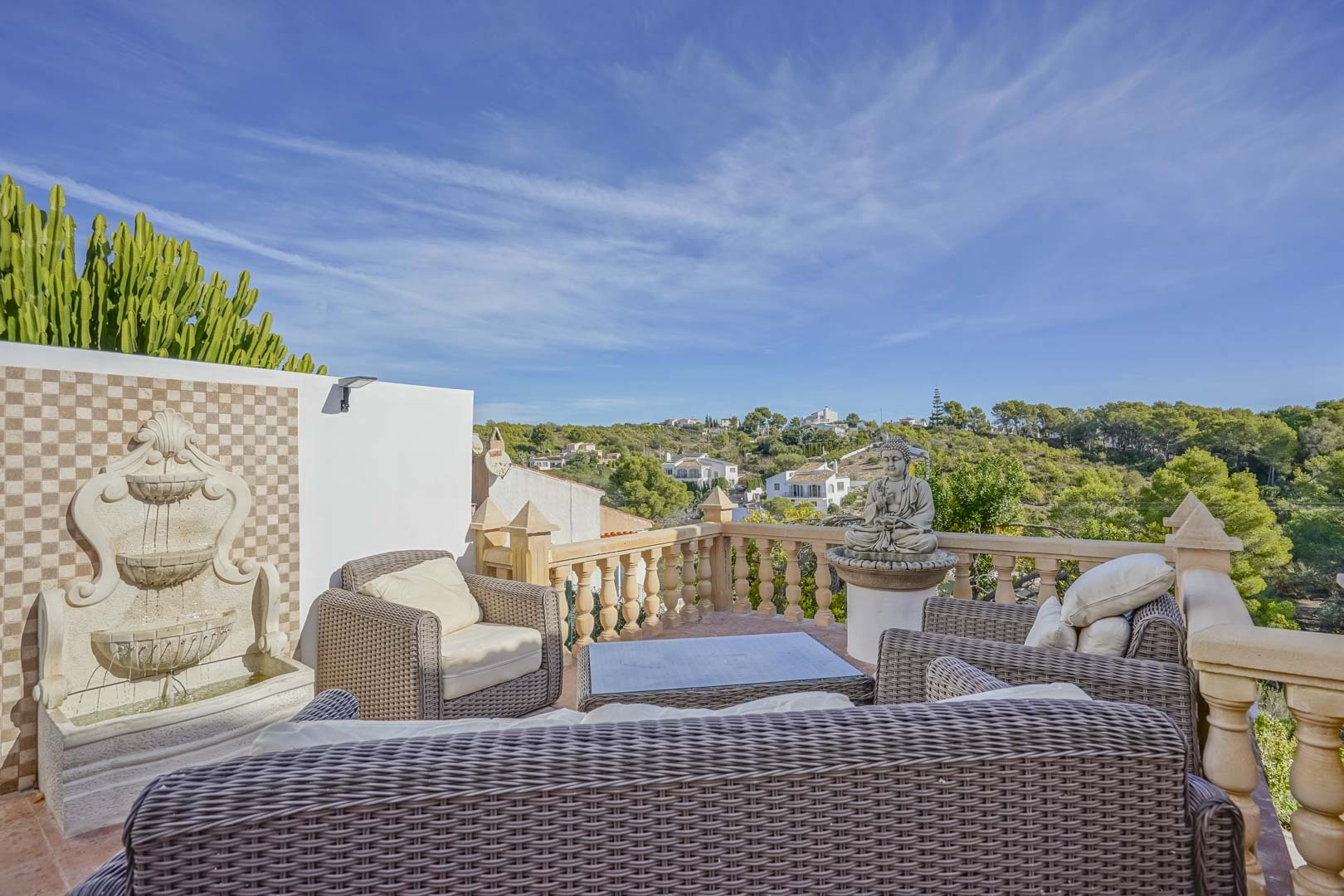 Elevated terrace featuring wicker lounge furniture, a tiered wall fountain, and stone balustrades overlooking the lush landscape of Jávea.
