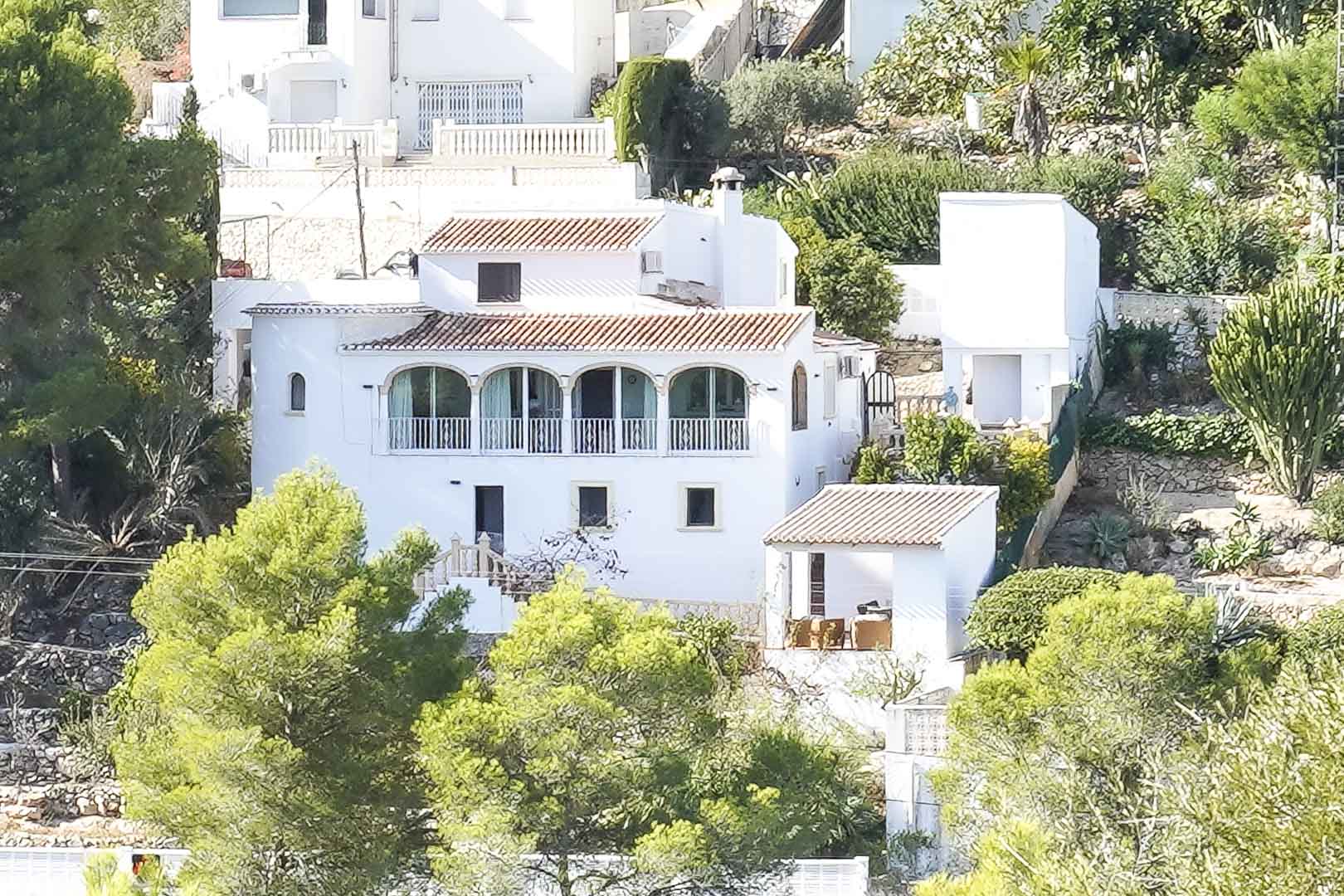 Exterior view of the white villa featuring traditional terracotta roofs and an arched naya, set within a sloped Mediterranean landscape with mature pines.