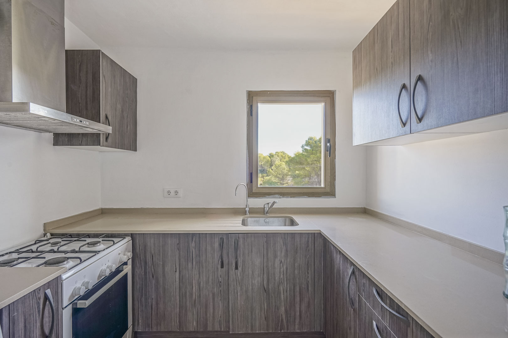 Functional kitchen featuring dark wood-effect cabinetry, neutral countertops, and a window overlooking greenery. Equipped with a gas stove and stainless steel extractor hood.