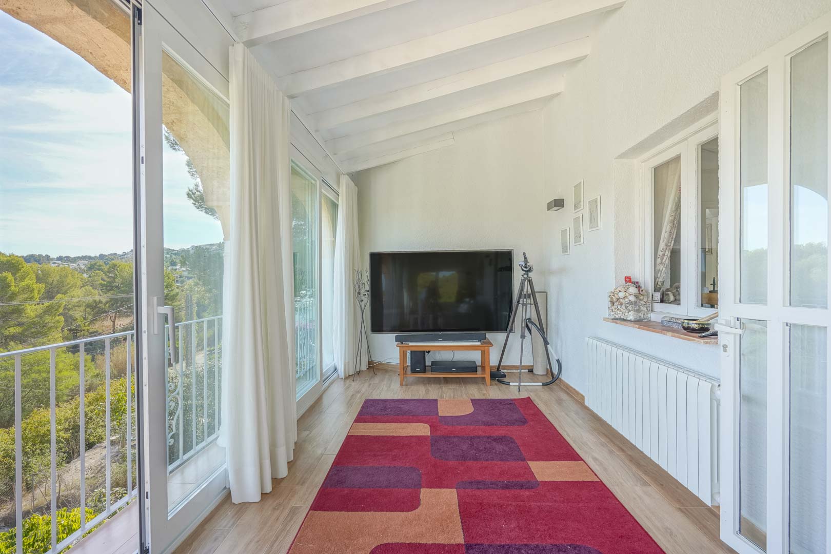 Glazed naya featuring white beamed ceilings, wood-effect flooring, and large sliding glass doors that open to scenic views of the surrounding greenery.