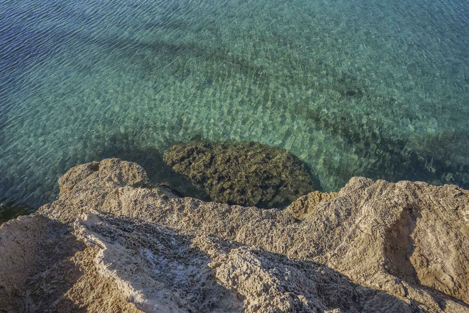 High-angle view of the rocky coastline in Jávea, featuring transparent turquoise waters and visible underwater rock formations.