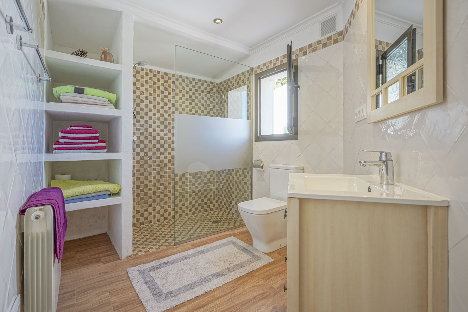 Renovated bathroom featuring a walk-in shower with mosaic tiling, built-in masonry shelving, wood-effect flooring, and a light wood vanity with a white ceramic sink.