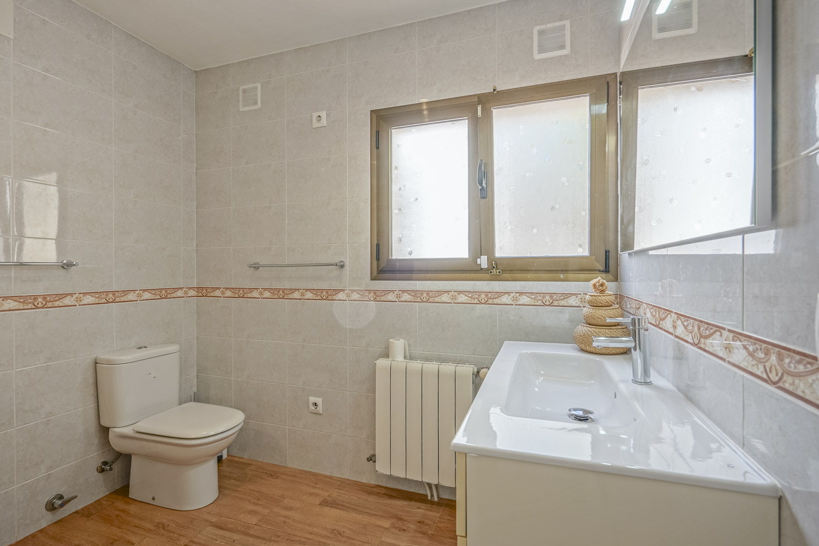Renovated bathroom featuring wood-effect flooring, tiled walls with a decorative border, a white vanity unit, and a radiator under a frosted window.