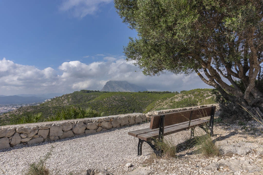 Scenic viewpoint with a wooden bench and dry-stone wall, offering panoramic views of the Montgó mountain and the surrounding green valleys.