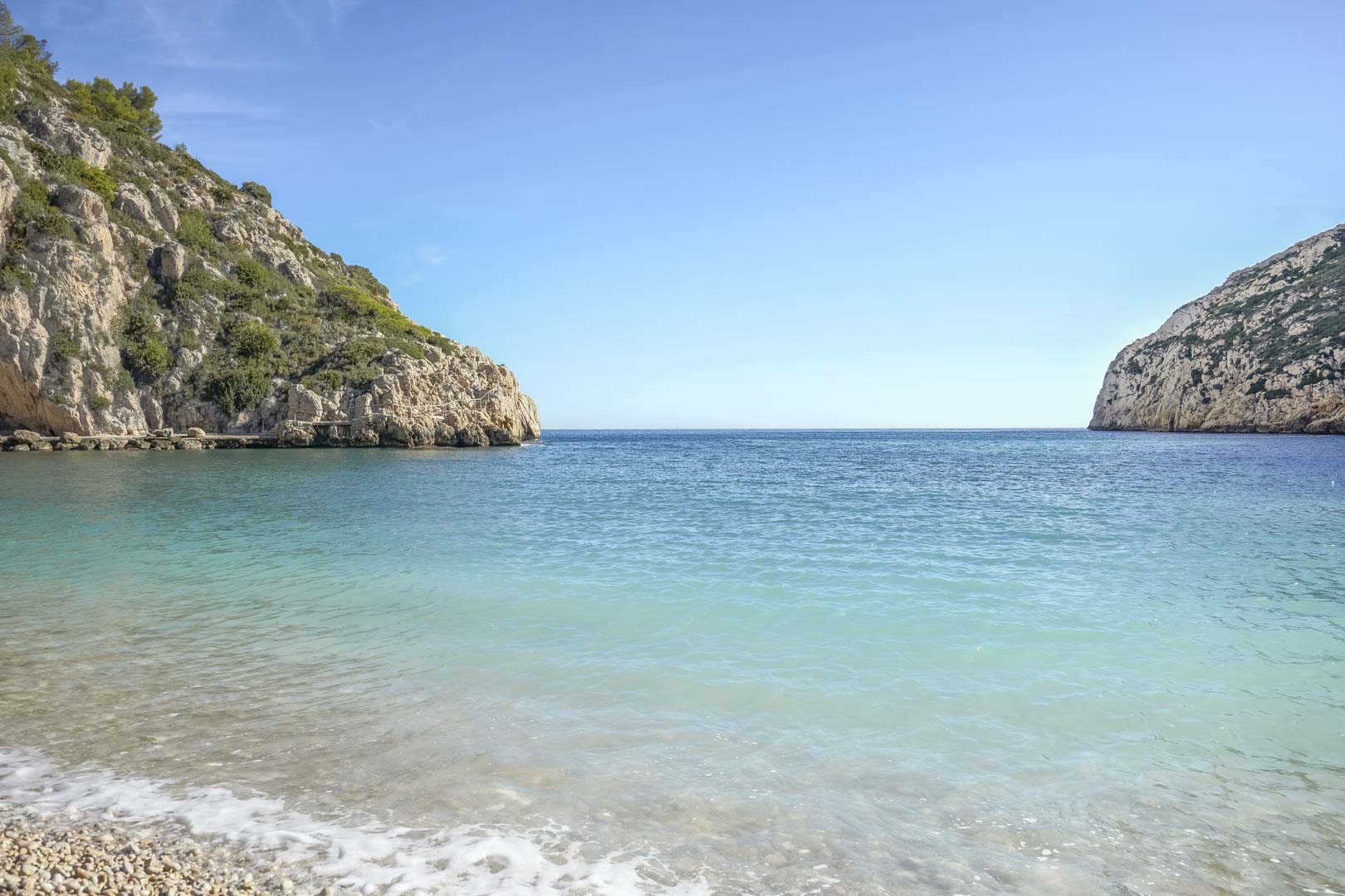 View of a Mediterranean cove with turquoise waters and a pebble shoreline, flanked by limestone cliffs under a clear sky near the property in Jávea.