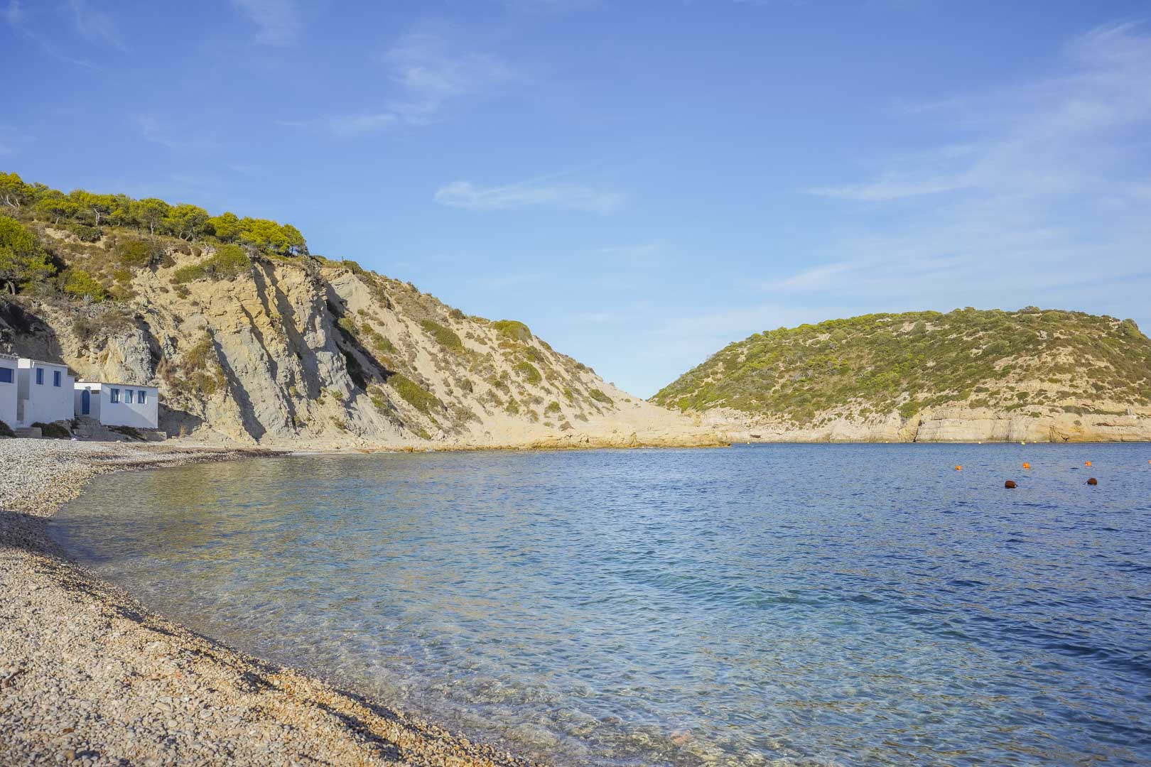 View of a pebble beach in Jávea featuring crystal clear waters, surrounded by rocky cliffs and Mediterranean vegetation under a bright blue sky.
