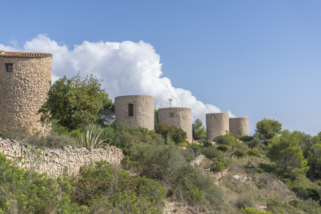 View of the historic stone windmills in Jávea, set on a hillside with native Mediterranean vegetation, dry stone walls, and clear blue skies.