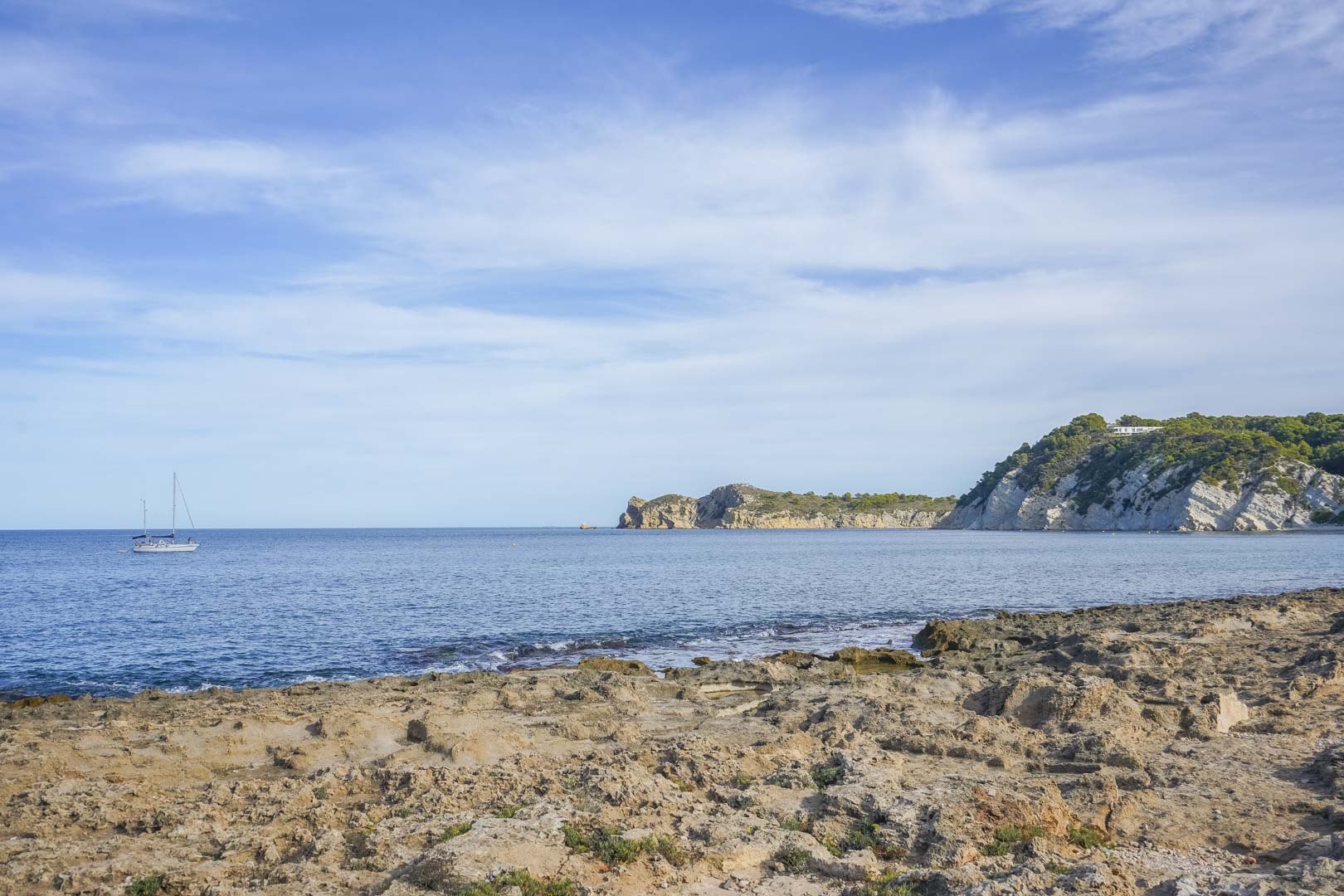 View of the rocky coastline in Jávea featuring the Mediterranean Sea, limestone cliffs, and a sailboat under a clear blue sky.