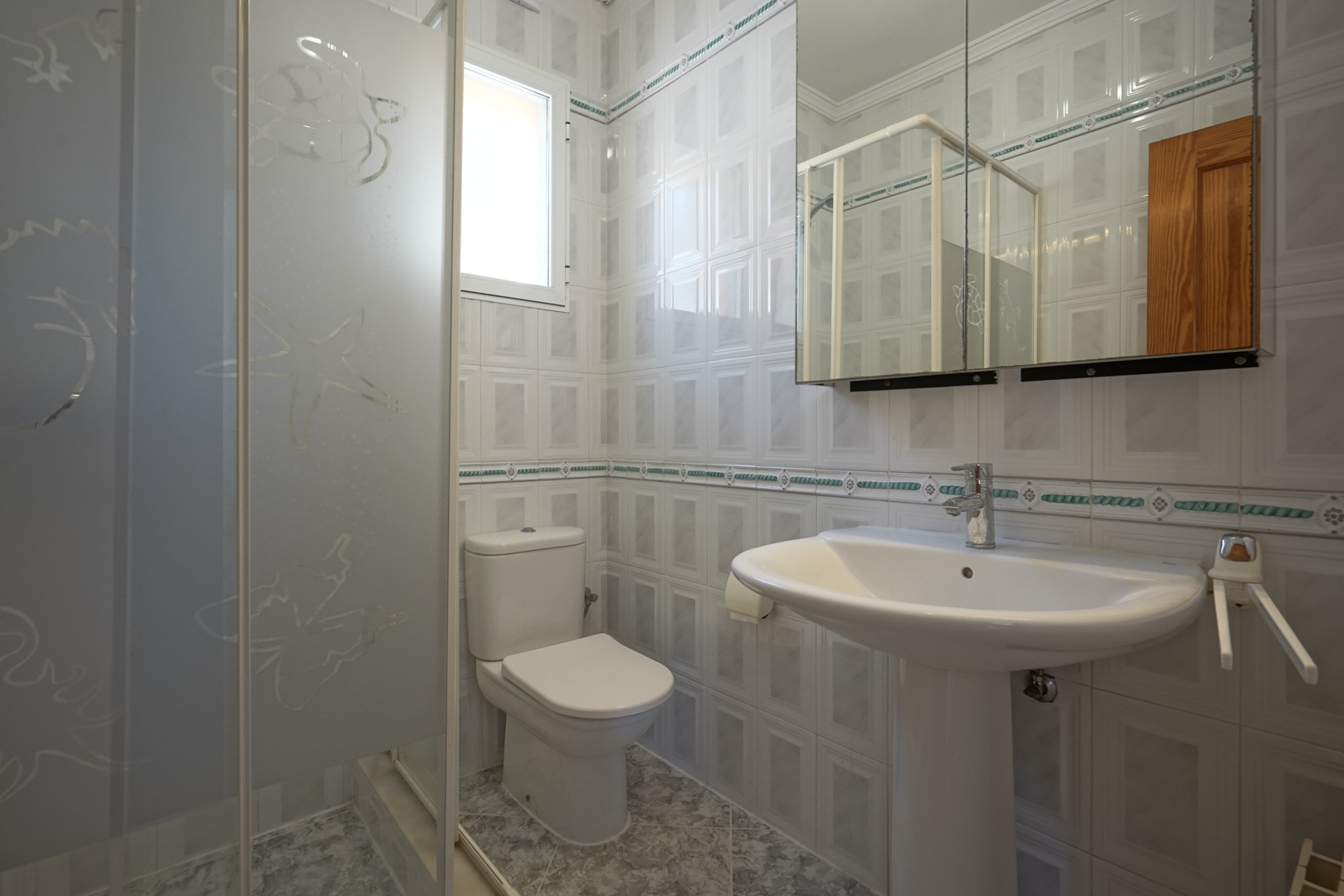 Bathroom featuring a pedestal sink, toilet, and walk-in shower with frosted glass panels. The space includes tiled walls with decorative borders and a mirrored cabinet.