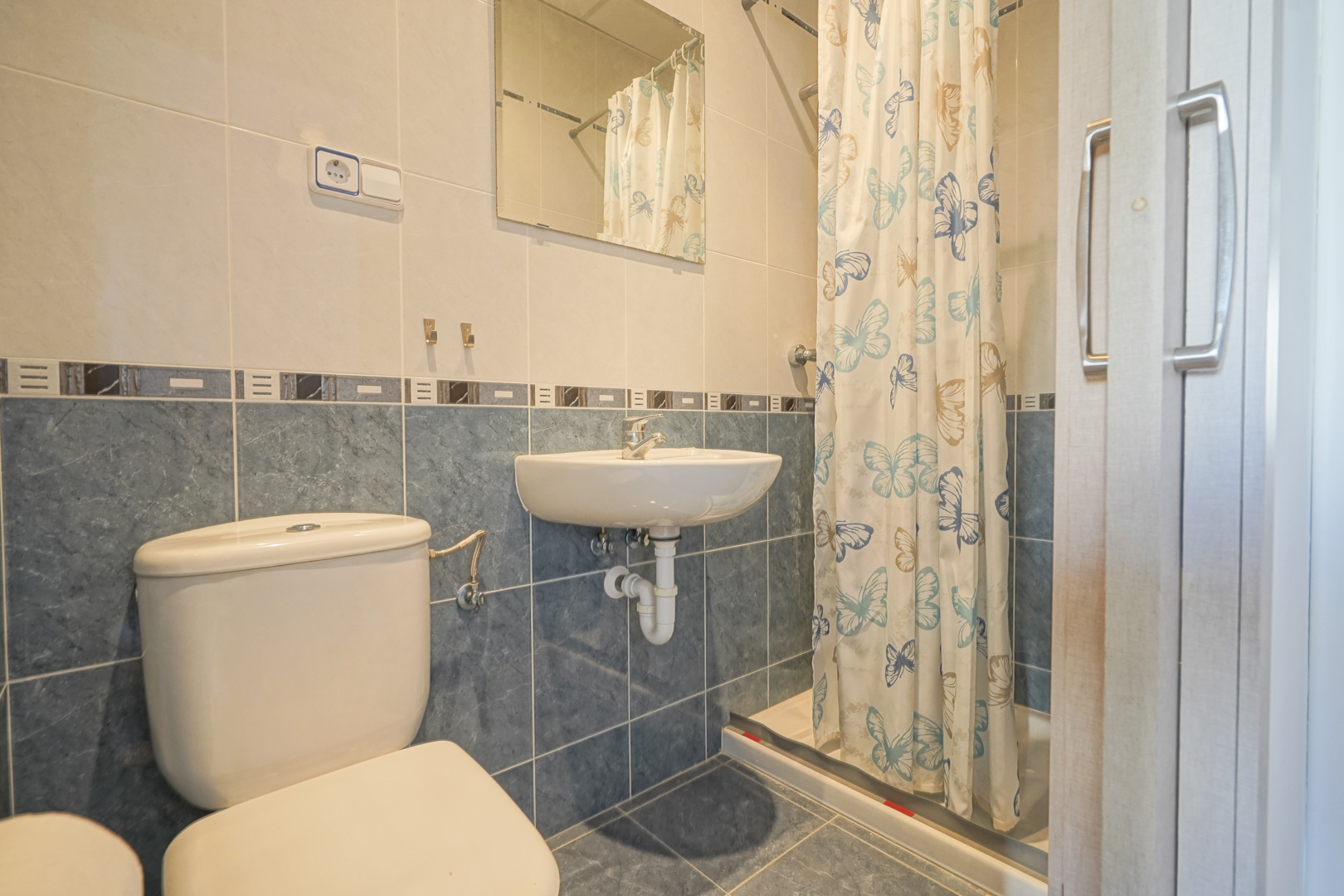 Bathroom featuring blue and cream tiling, a wall-mounted washbasin, toilet, and a shower area enclosed by a butterfly-patterned curtain.