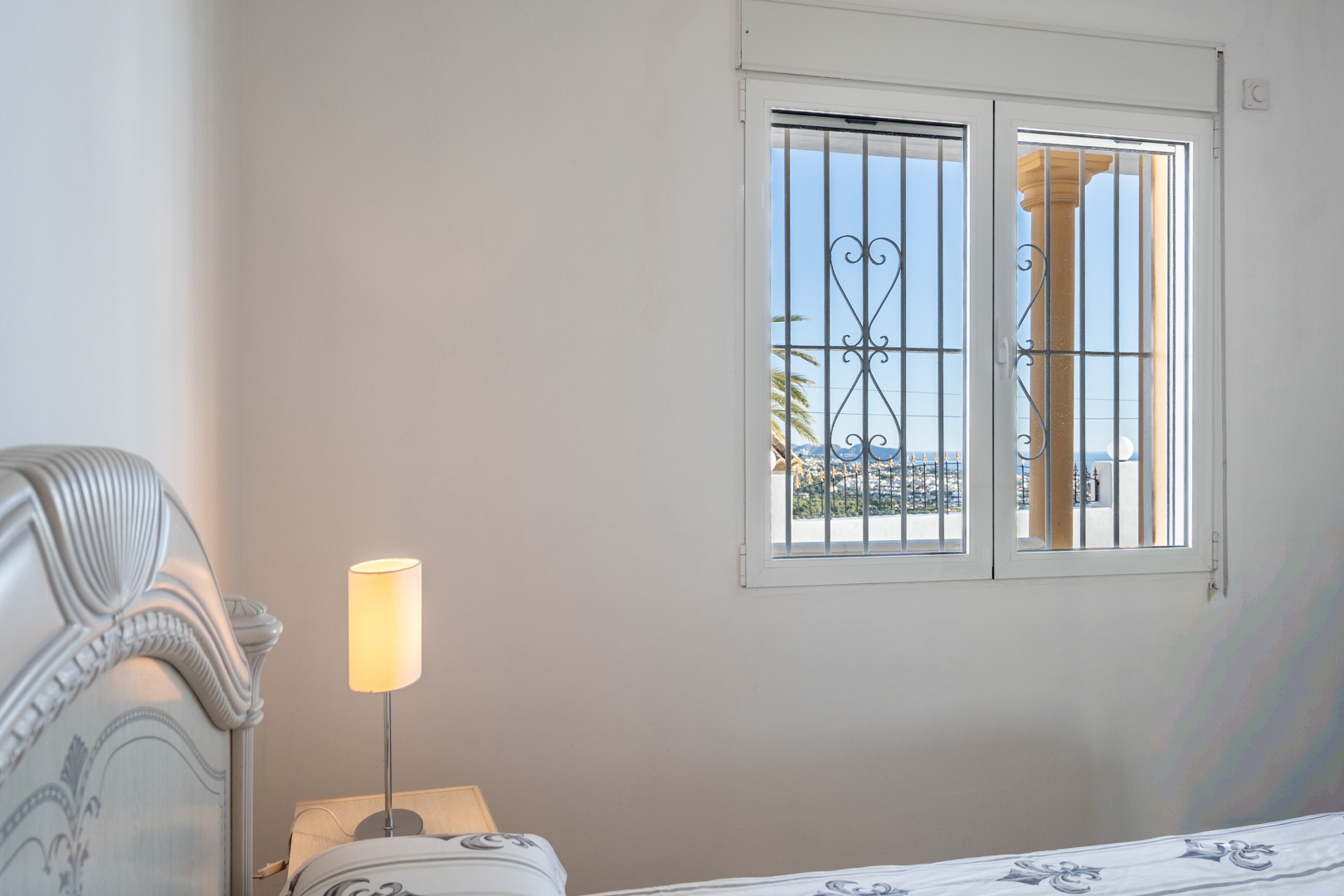 Bedroom featuring a classic white headboard and a window with decorative security bars, providing views of the Calpe skyline and sea.