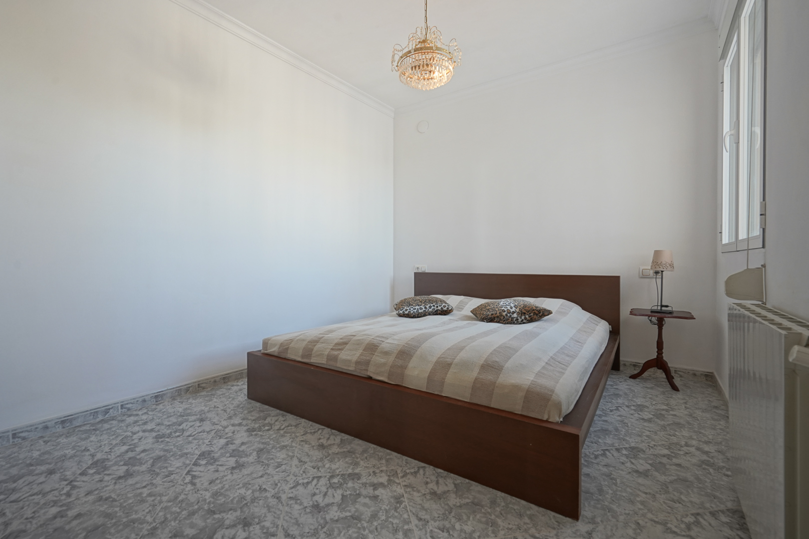 Bedroom featuring a dark wood bed frame, marbled tile flooring, a classic crystal chandelier, and natural light from a side window.