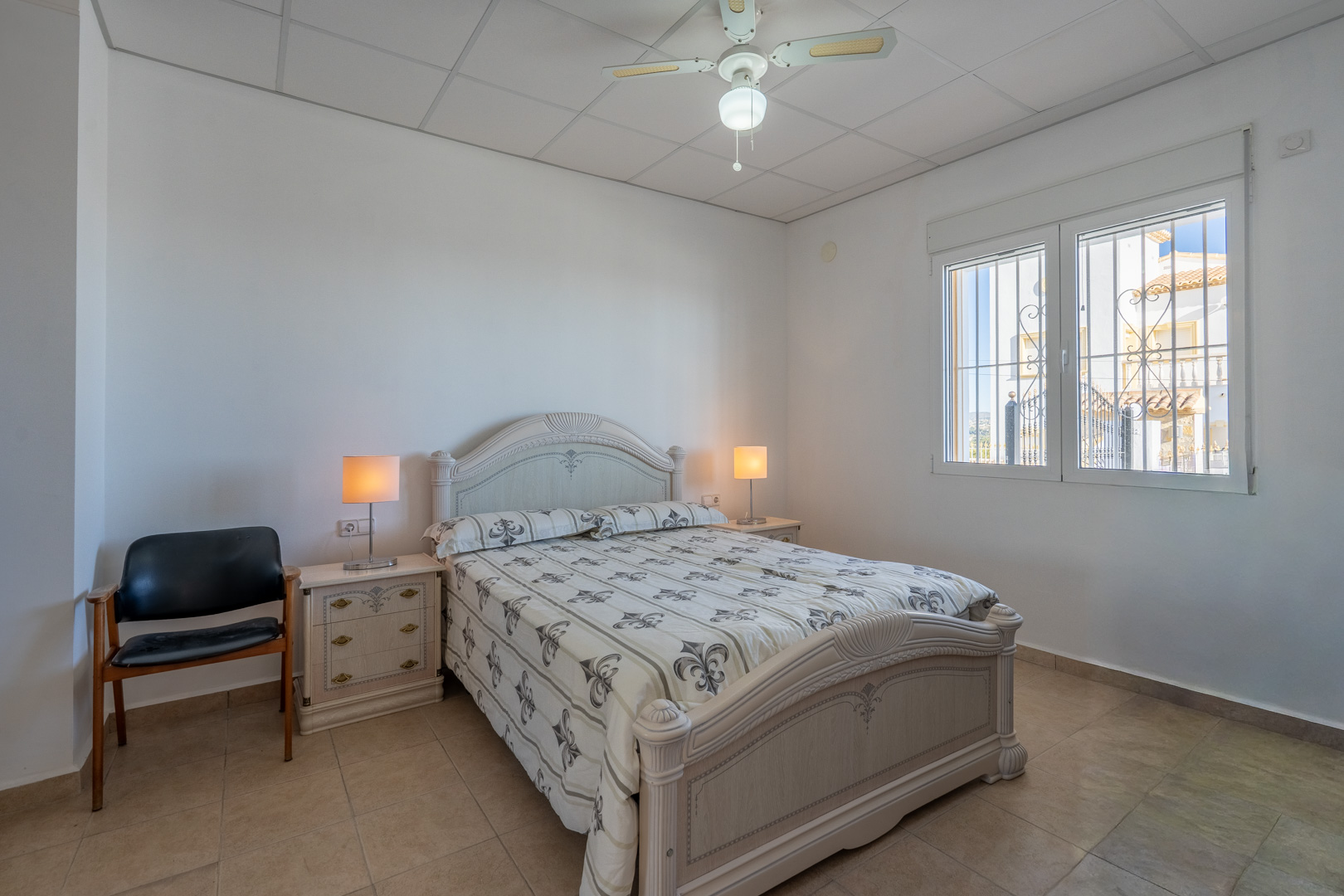 Bedroom featuring traditional wooden furniture, tiled flooring, a ceiling fan, and a window with security bars providing natural light.