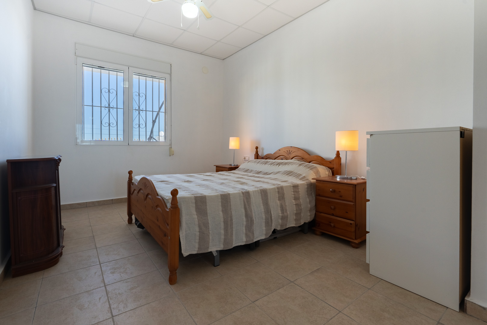 Bedroom featuring traditional wooden furniture, tiled flooring, and a window equipped with security bars and a roller shutter for natural light.