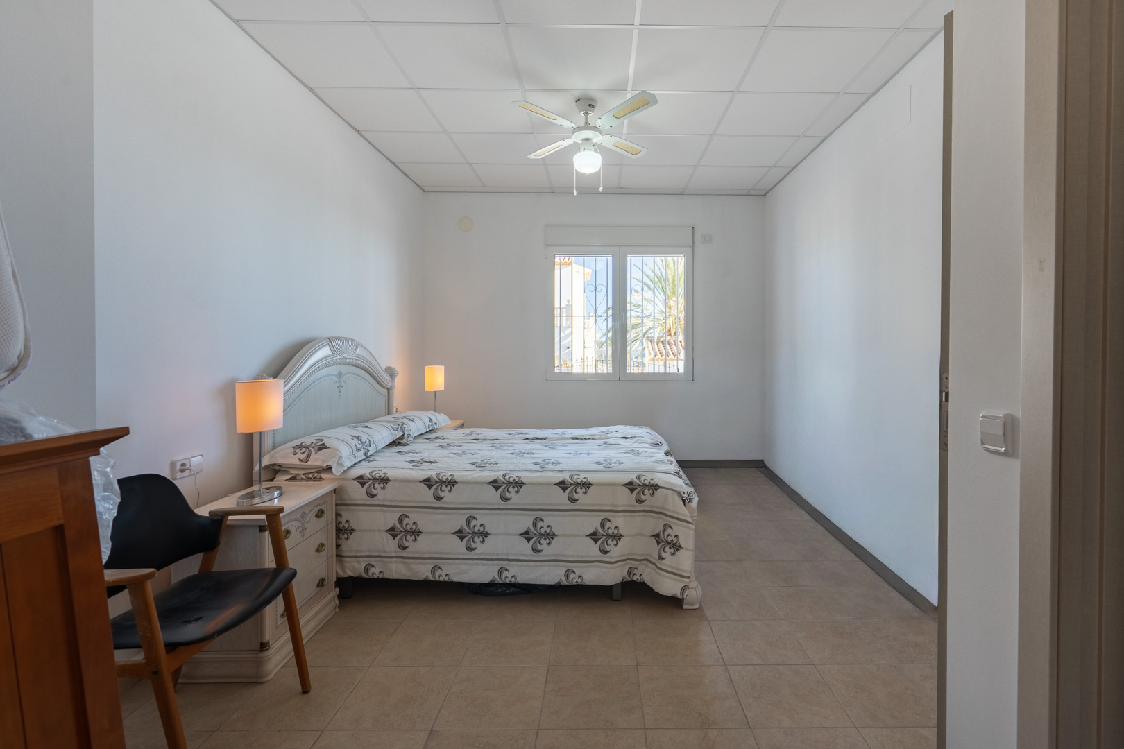 Bright bedroom featuring a double bed with an ornate headboard, ceiling fan, and tiled flooring. A window offers natural light and views of the surrounding palm trees.
