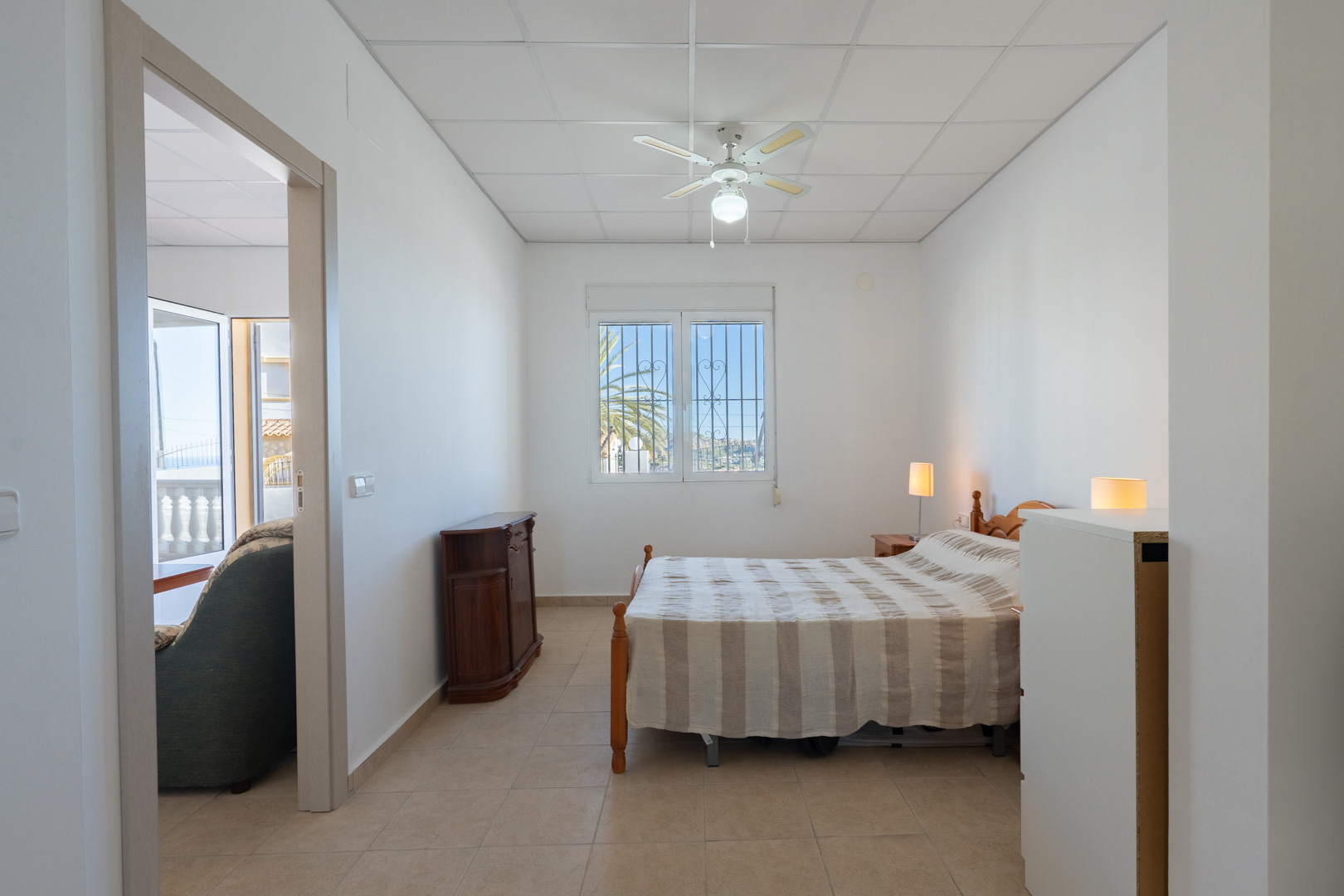 Bright bedroom featuring ceramic tile flooring, a ceiling fan, and a window with security bars. The room connects to the living area and offers views of the surrounding palm trees.