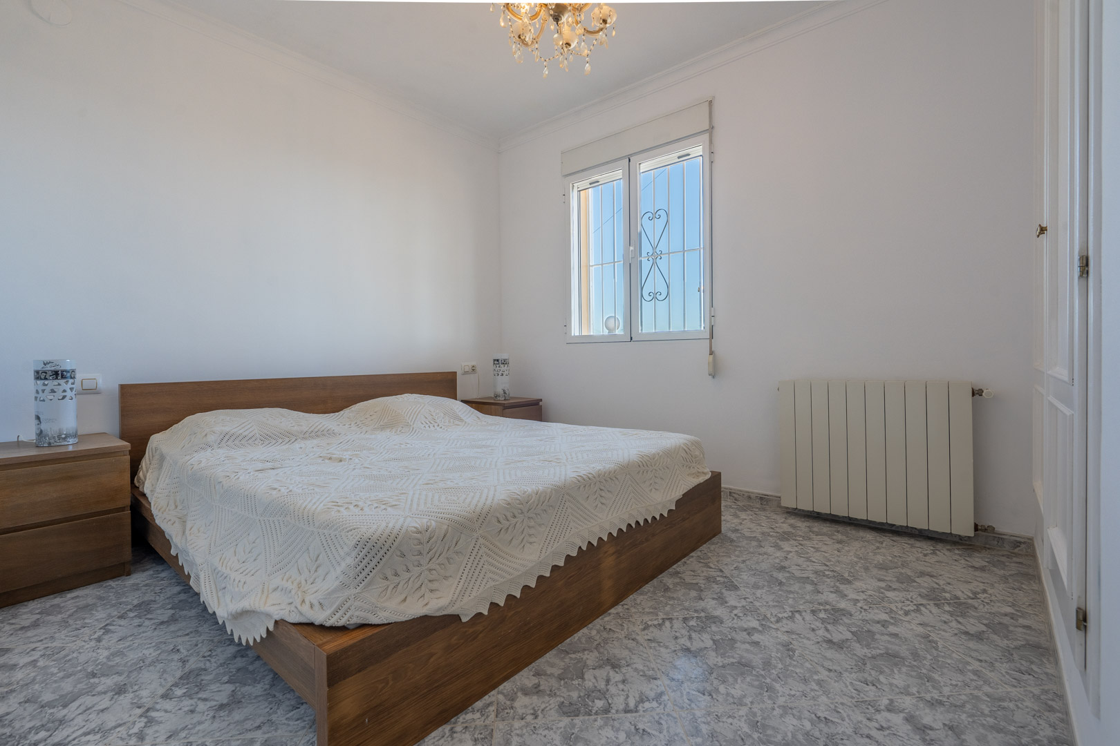 Bright bedroom featuring marble-effect tiled flooring, wooden bed frame with matching nightstands, built-in white wardrobes, and central heating radiator.