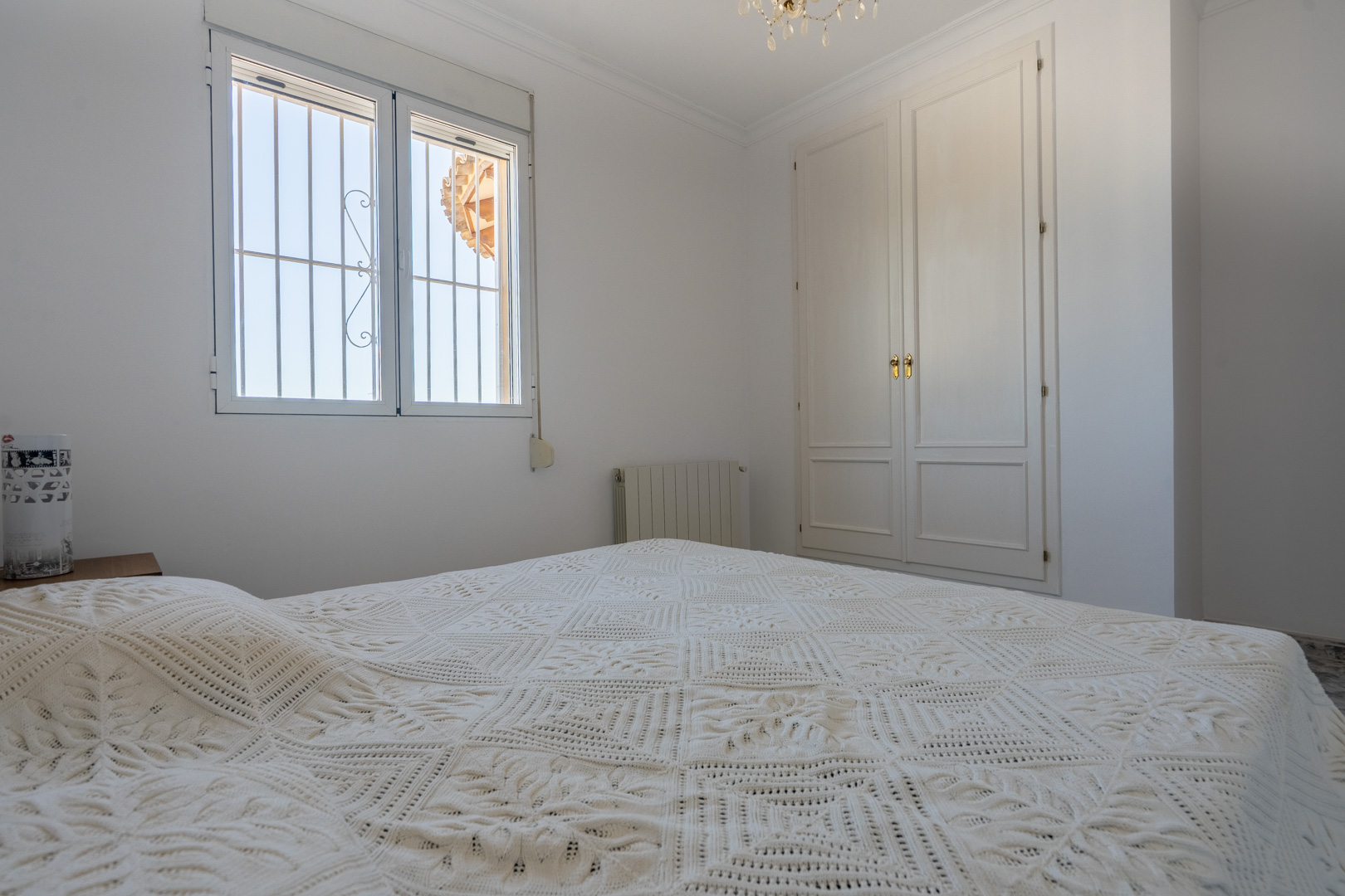 Bright bedroom featuring white walls, a built-in wooden wardrobe, and a window with security bars. The space includes a radiator and ample natural lighting.