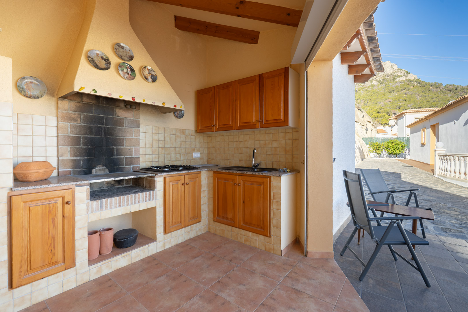 Covered summer kitchen featuring a built-in brick barbecue, wooden cabinetry, granite countertops, and a sink, adjacent to a terrace with mountain views.