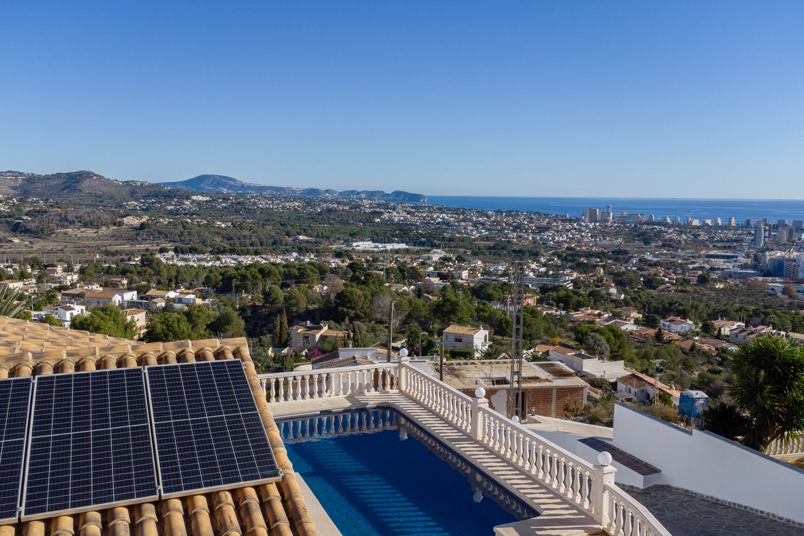 Elevated view featuring a swimming pool with classic balustrades, rooftop solar panels, and a wide panorama of the Calpe coastline and Mediterranean Sea.