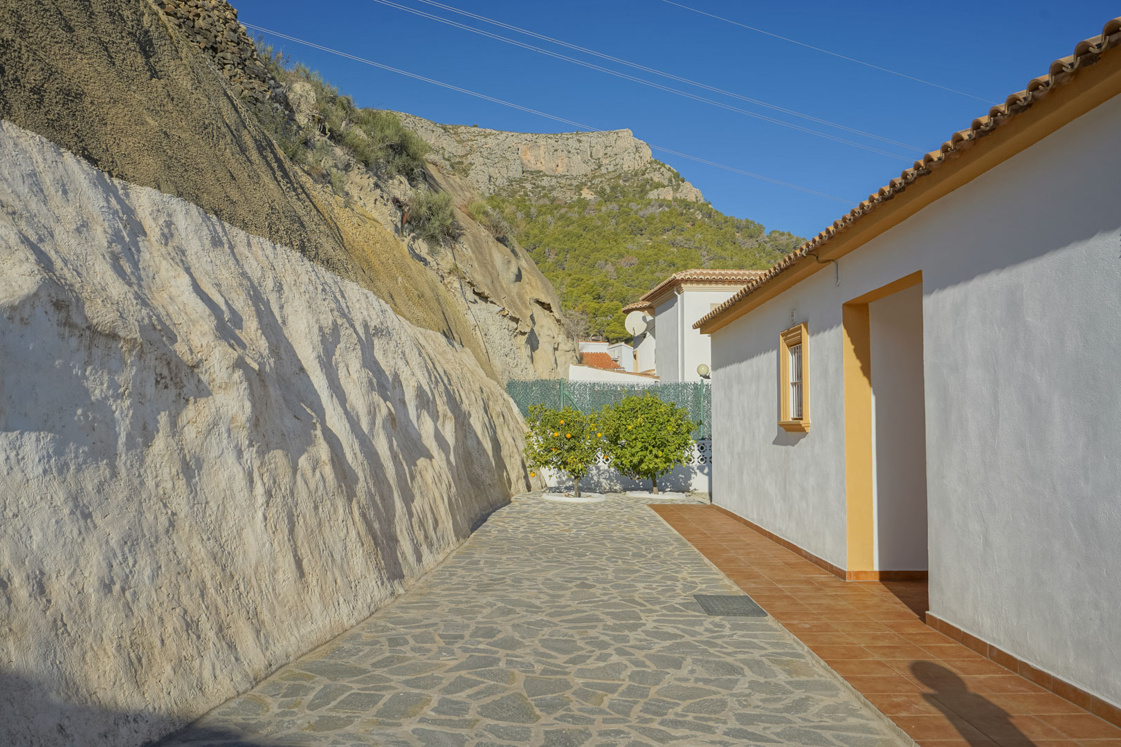 Exterior stone-paved walkway set against a natural rock face, with views of the Sierra de Oltà mountains and fruiting citrus trees.