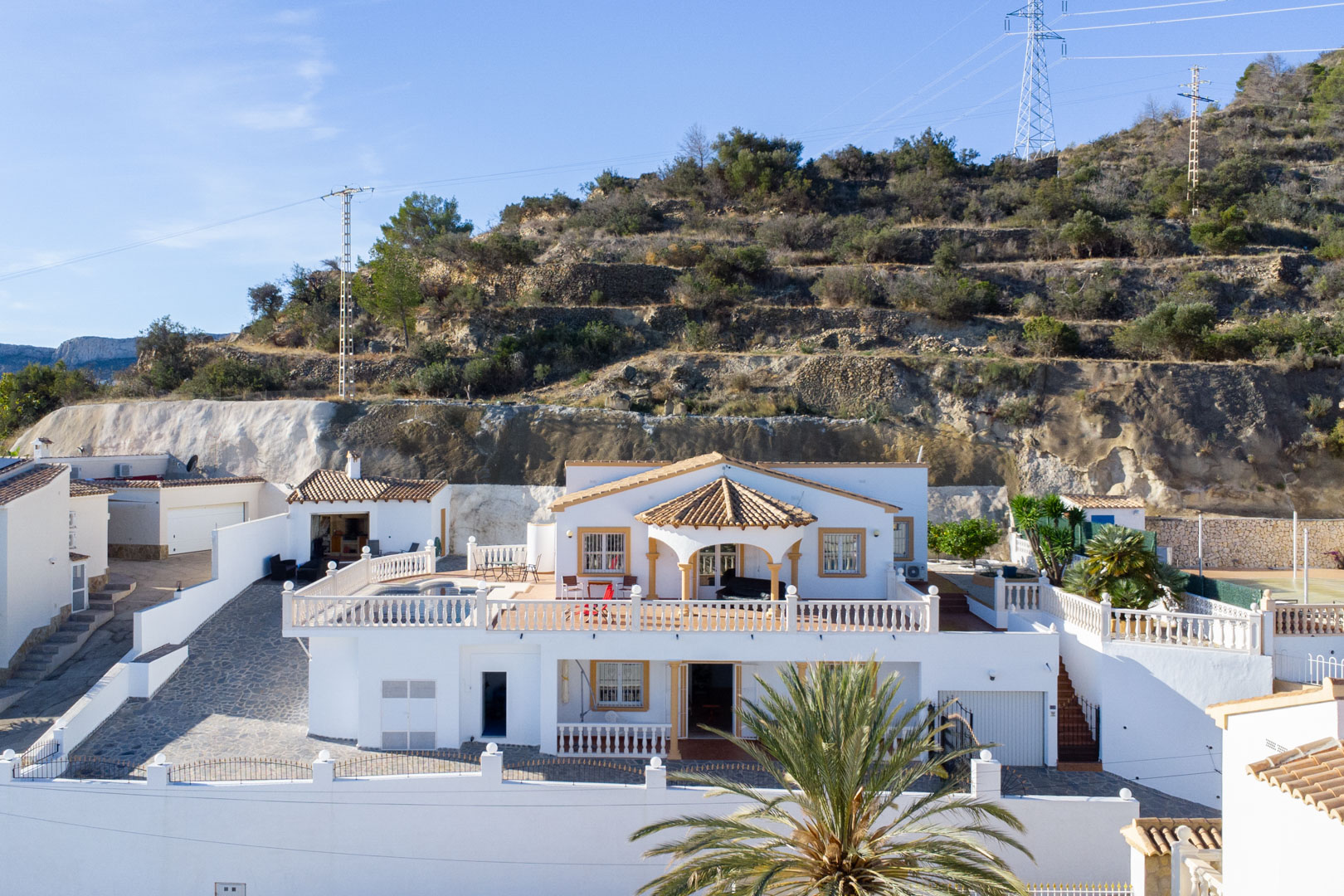 Exterior view of the two-level white villa featuring expansive balustraded terraces, a private pool, and a summer kitchen, set against the Sierra de Oltà.