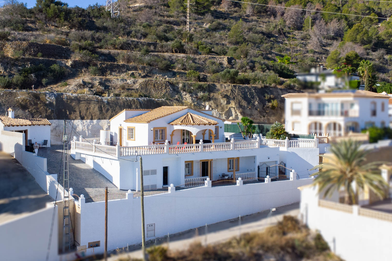 Exterior view of the two-story villa featuring a white facade, terracotta roof, and expansive terraces with stone balustrades set against the Sierra de Oltà hillside.