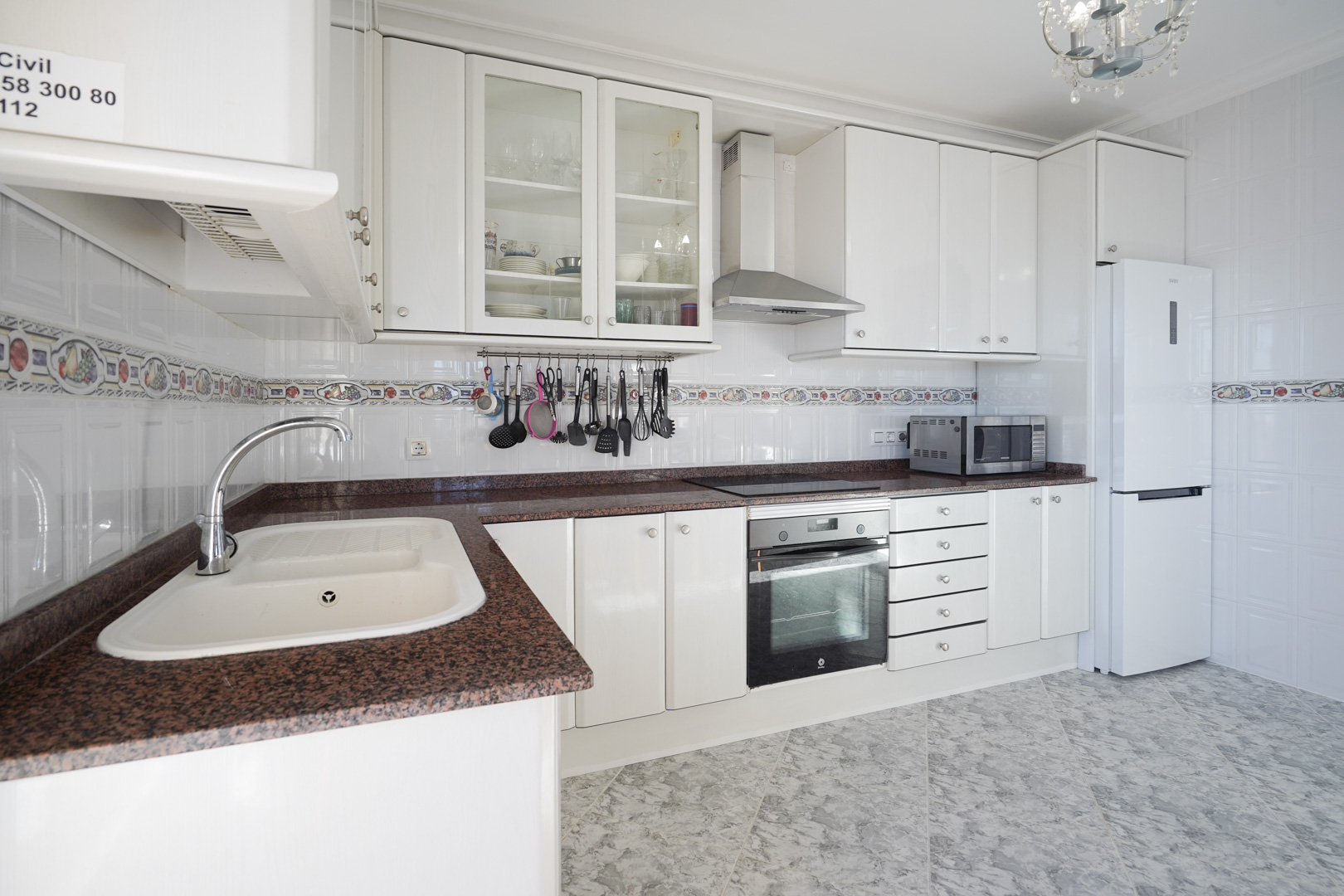 Fully equipped kitchen featuring white cabinetry, reddish granite countertops, and tiled walls with a decorative border. It includes integrated appliances and marble-effect flooring.