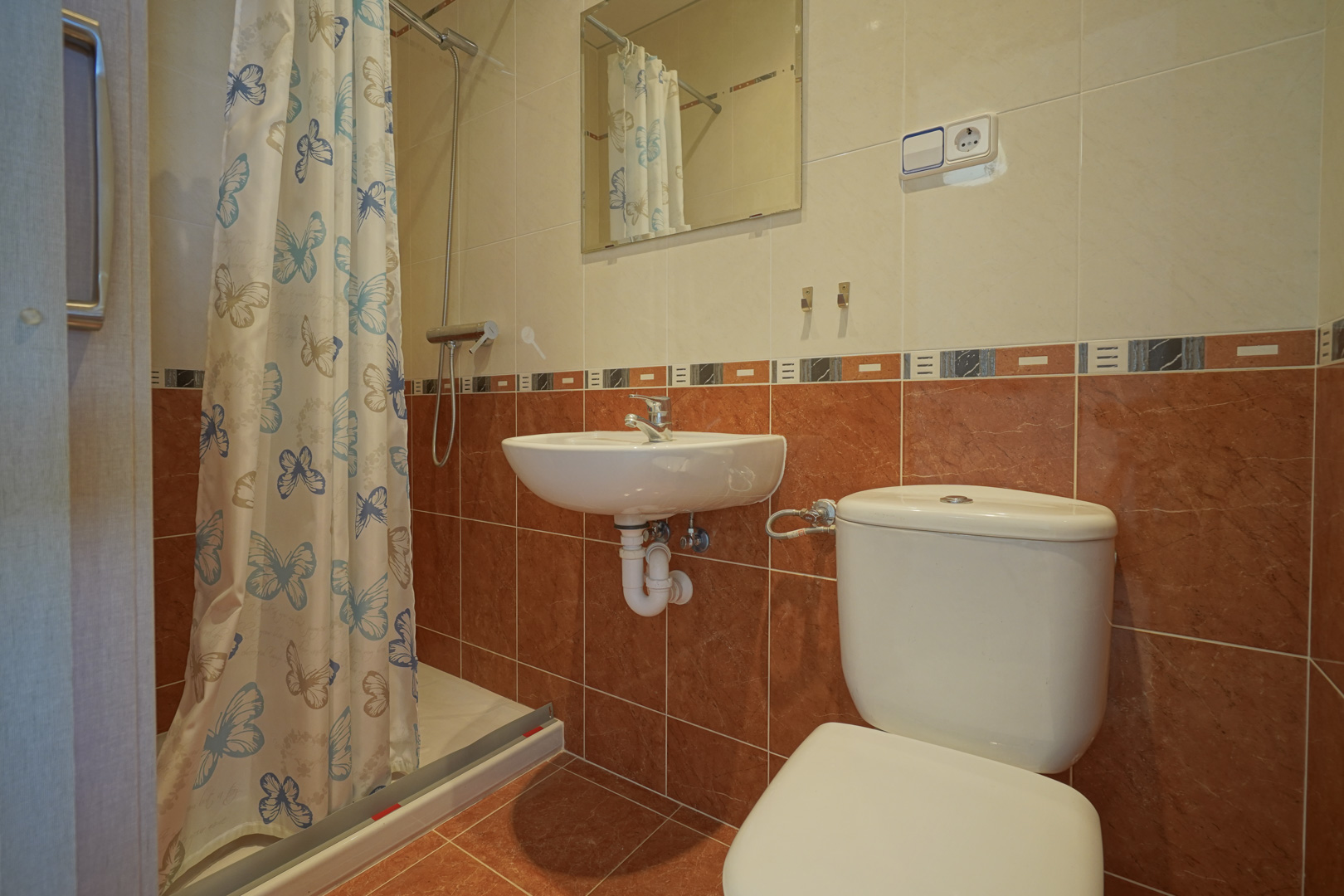Functional bathroom featuring a walk-in shower with butterfly curtain, wall-mounted sink, and terracotta and cream wall tiling.