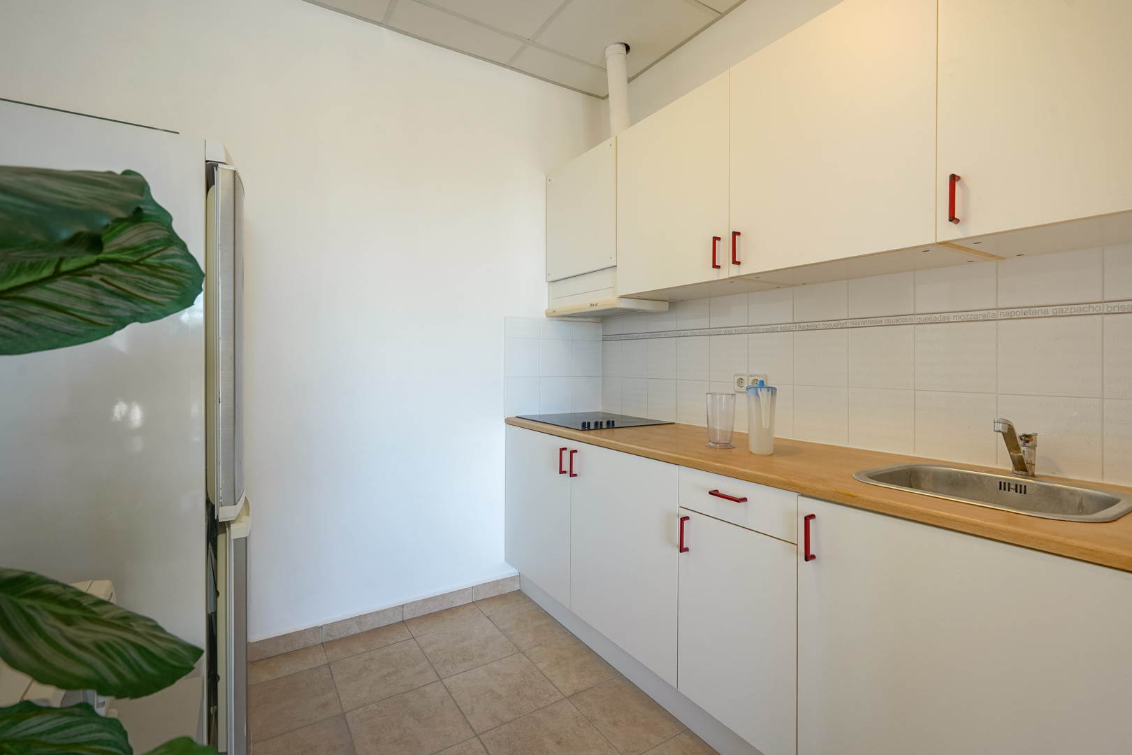 Functional kitchen featuring white cabinetry with red handles, a wooden countertop, and integrated appliances including an electric cooktop and stainless steel sink.