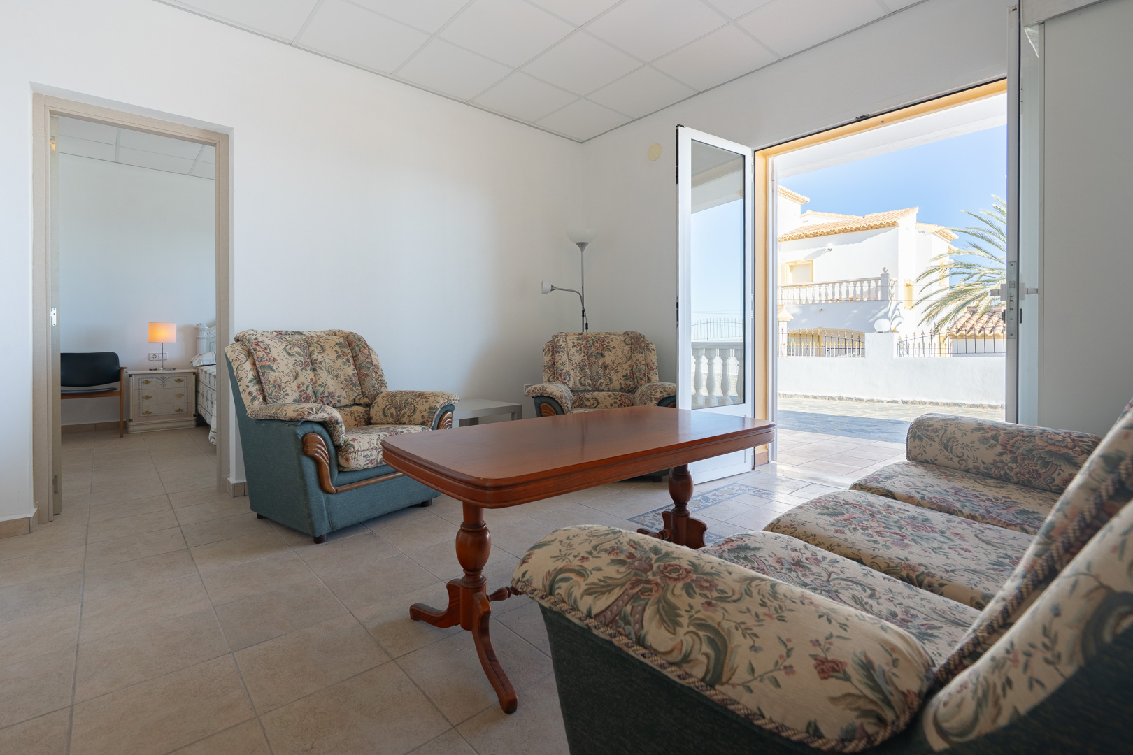 Guest apartment living area featuring floral upholstery, a wooden coffee table, and direct terrace access. The space includes tiled flooring and a view into a bedroom.