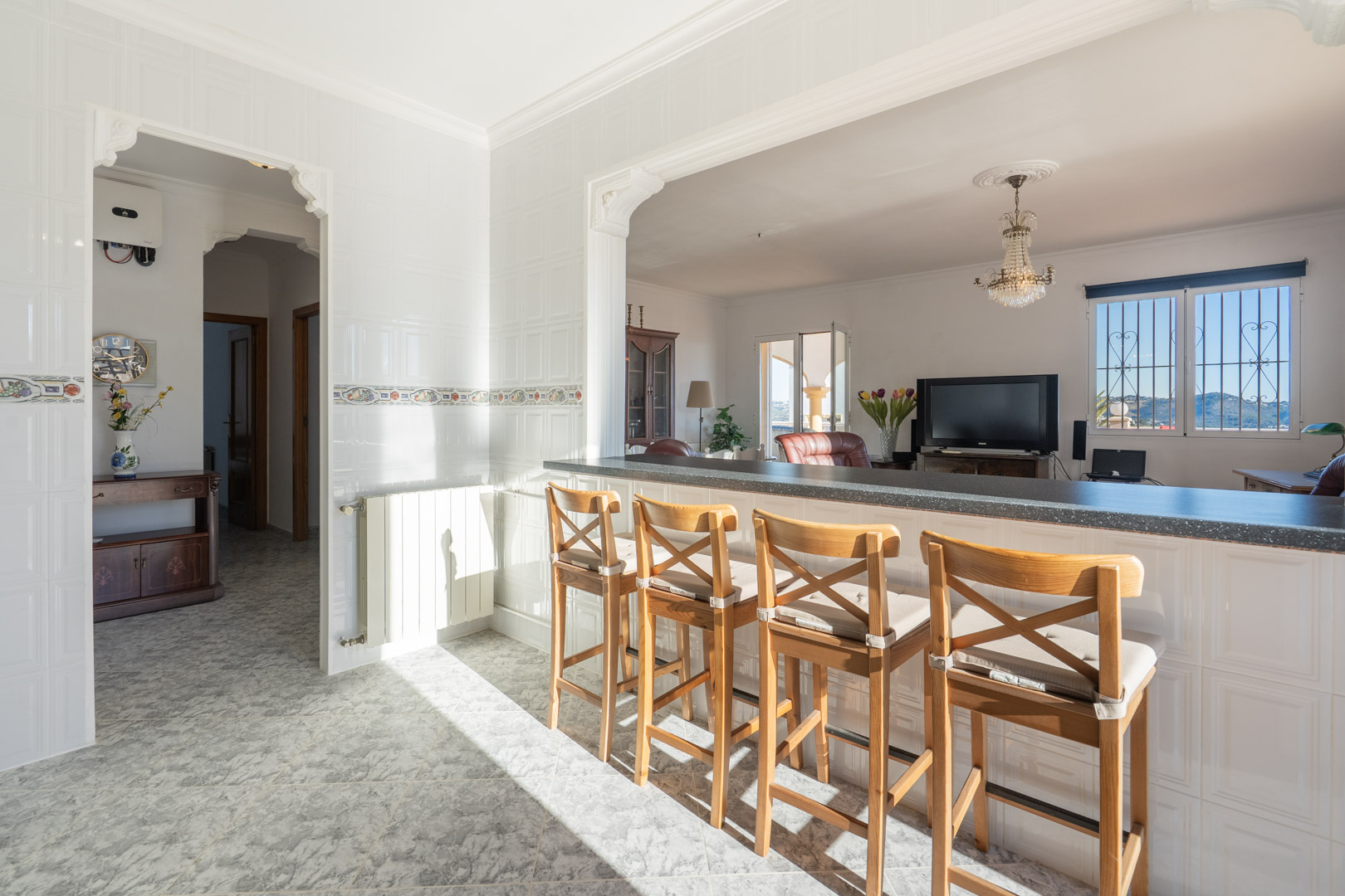 Kitchen breakfast bar with wooden stools overlooking the living area, featuring white tiled walls, traditional archways, and mountain views through the window.
