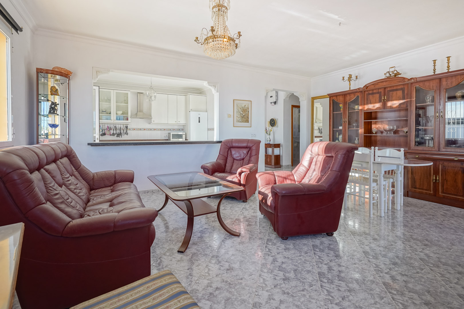 Living-dining area featuring marble-effect flooring, burgundy leather sofas, and a semi-open kitchen. Includes traditional wooden cabinetry and a crystal chandelier.
