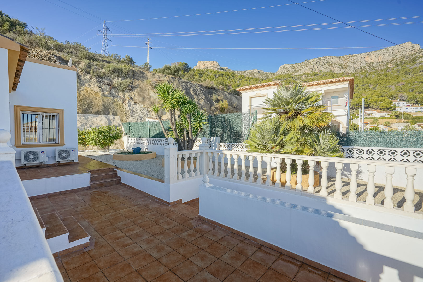 Outdoor terrace featuring rustic ceramic tiling and white balustrades, overlooking a gravel garden with palm trees against the backdrop of the Sierra de Oltà.