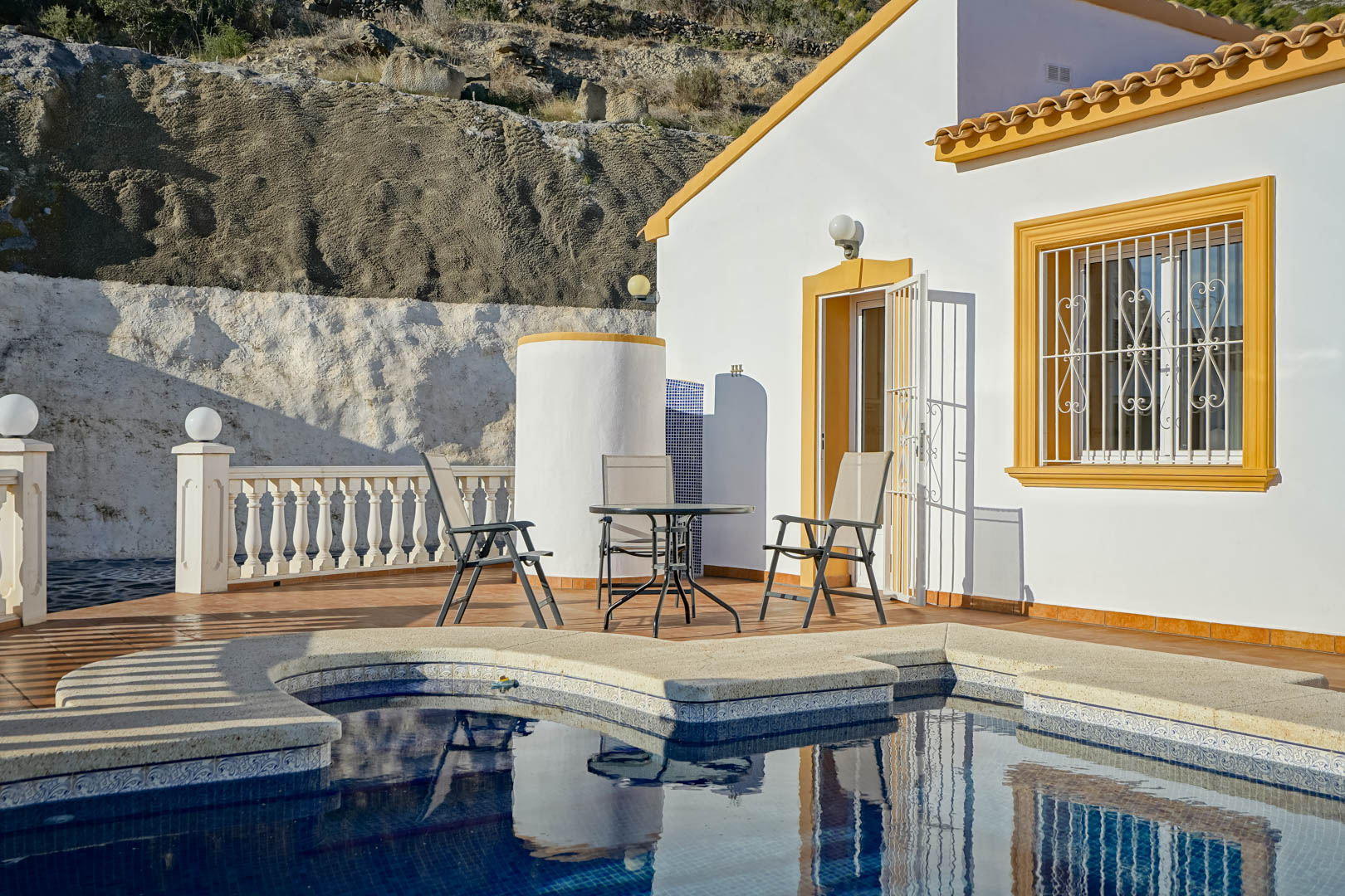 Poolside terrace featuring outdoor seating, a cylindrical shower, and a white villa facade with traditional ochre window frames against a rocky backdrop.