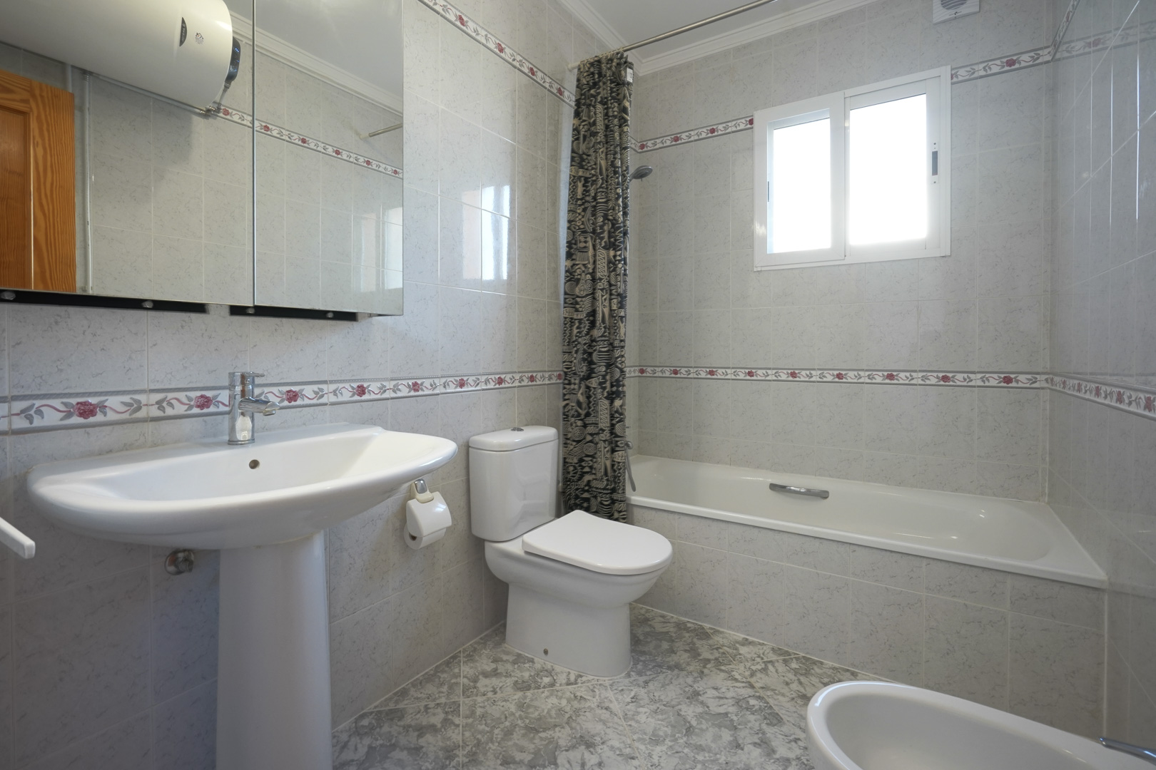 Traditional bathroom featuring tiled walls with floral borders, a bathtub with shower, pedestal sink, and a mirrored vanity cabinet.