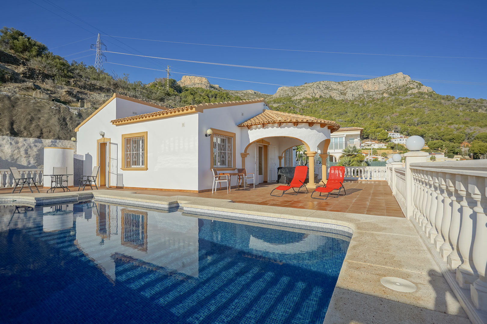 Villa exterior featuring a private swimming pool, terracotta terrace with sun loungers, and a traditional arched porch set against the Sierra de Oltà mountains.