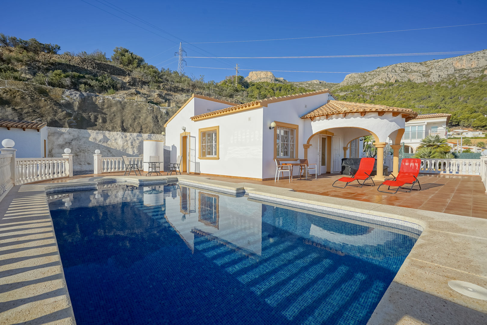 Villa exterior featuring a private swimming pool, terracotta terrace with sun loungers, and an arched porch set against the Sierra de Oltà mountain backdrop.