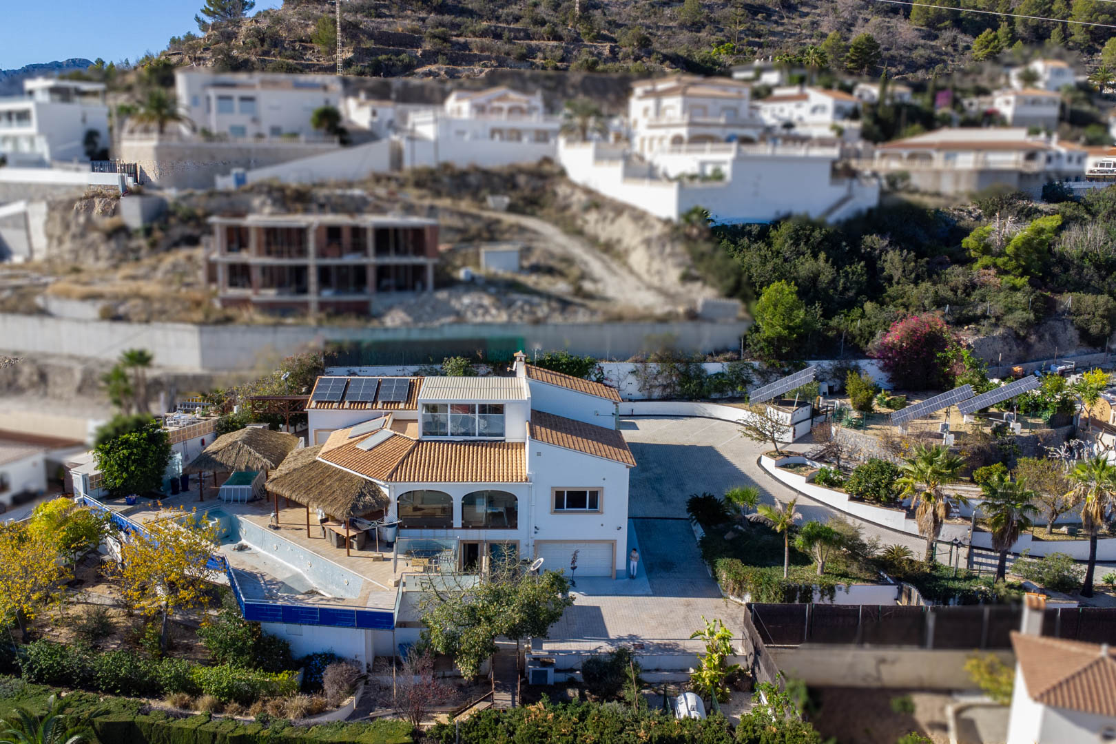 Aerial perspective of the villa featuring three motorized solar tracking towers, a spacious paved driveway, and outdoor terraces with thatched pergolas by the pool area.