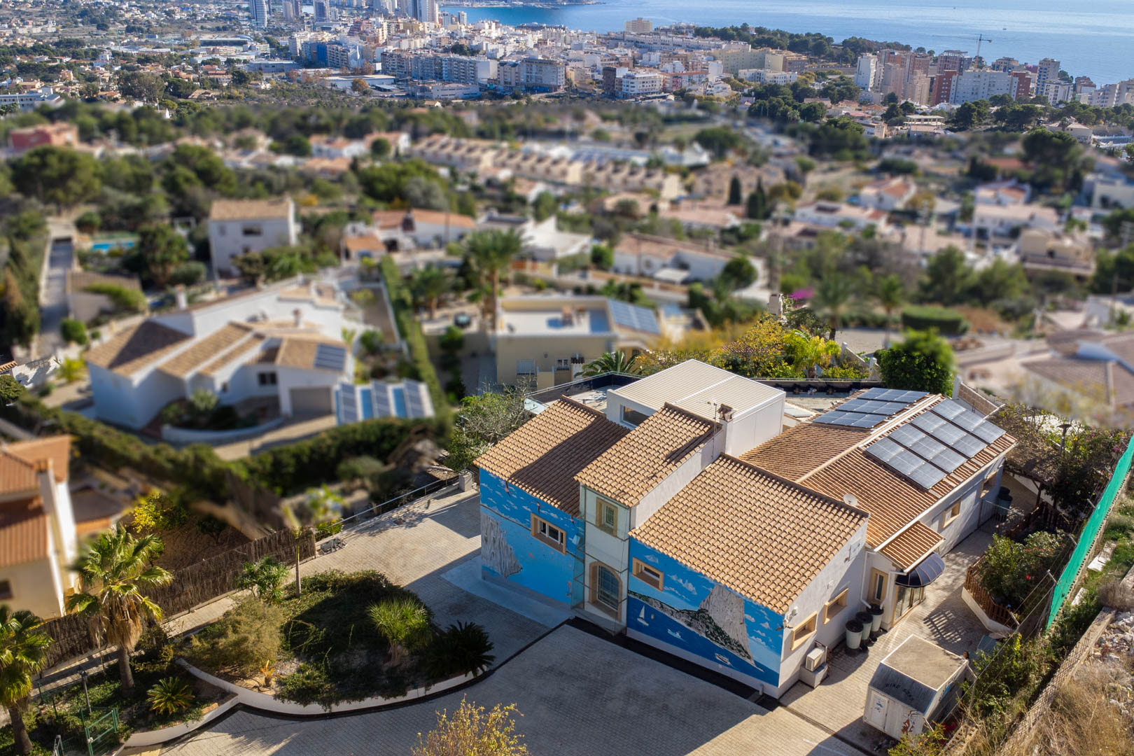 Aerial view of the villa featuring artistic murals, traditional tile roofing, and solar panels, set against the backdrop of Calpe's coastline and urban skyline.