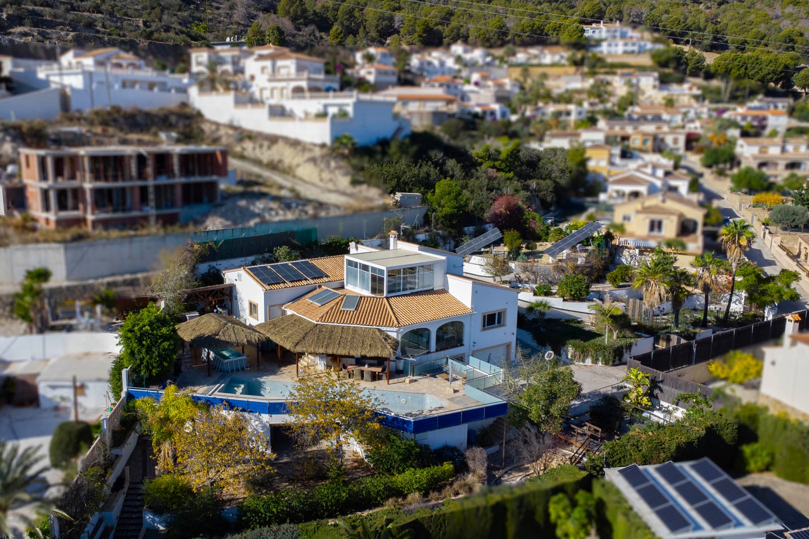 Aerial view of the villa featuring the pool area with thatched pergolas, rooftop solar panels, and motorized solar trackers within the landscaped grounds.