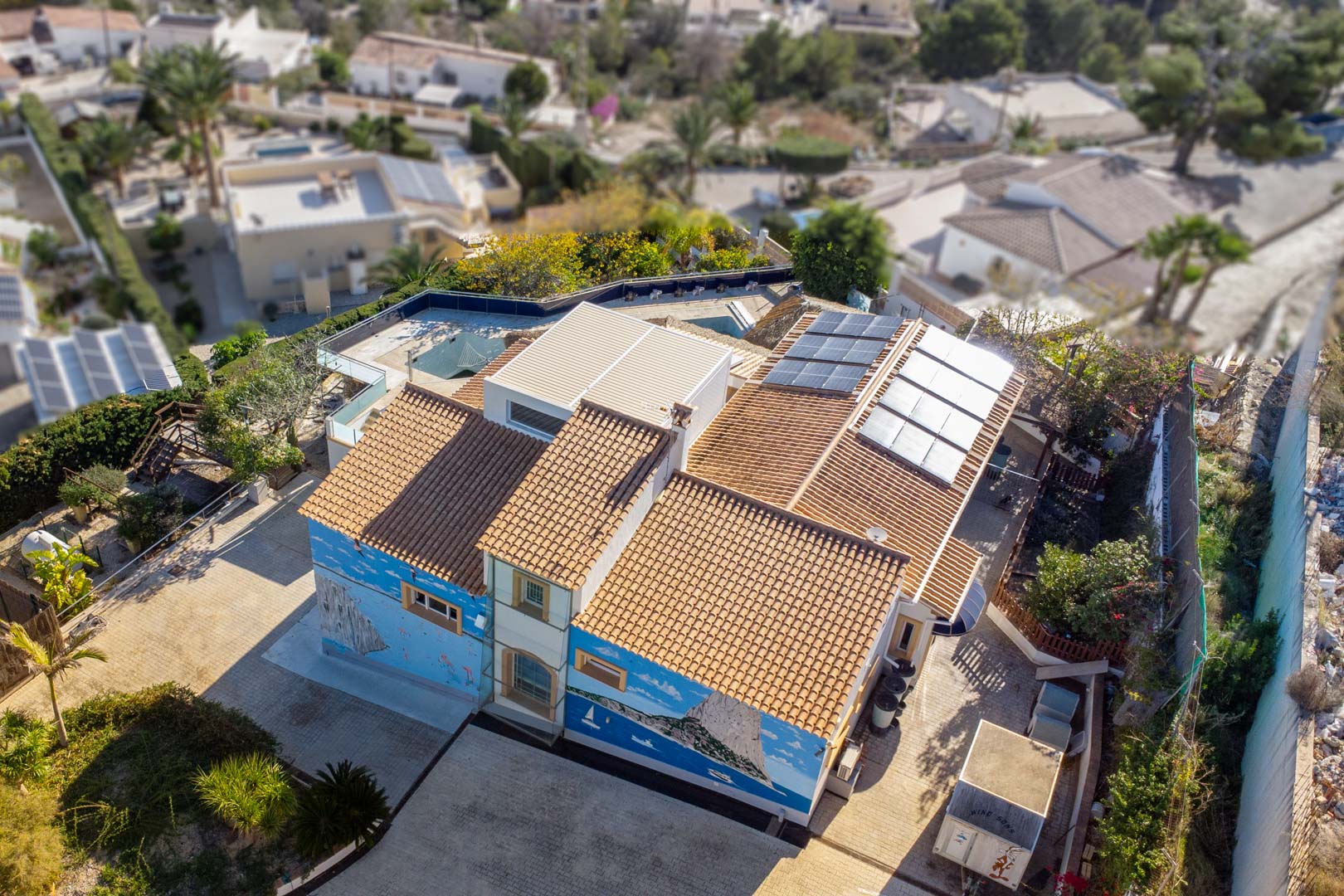 Aerial view of the villa featuring traditional tile roofing, integrated solar panels, and a large custom mural depicting the Peñón de Ifach on the exterior wall.