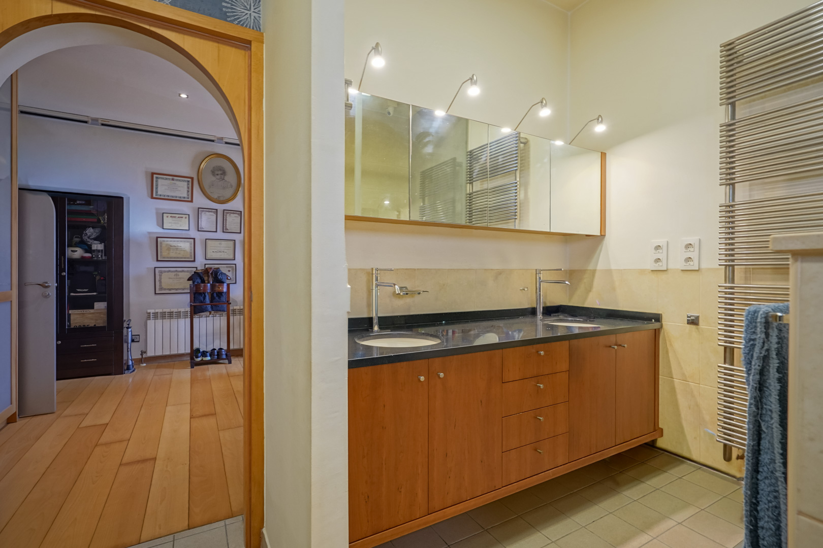 Bathroom featuring a double wooden vanity with a black countertop and illuminated mirrors. An arched doorway connects to a dressing area with hardwood flooring.
