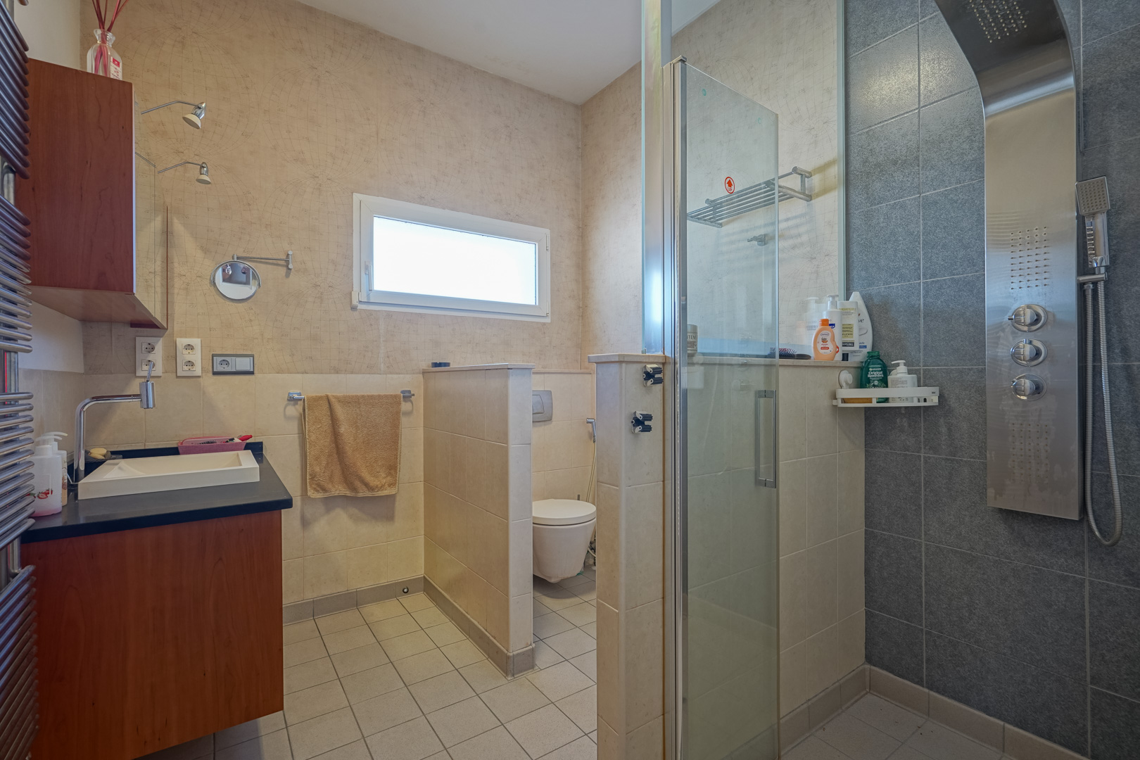 Bathroom featuring a hydromassage shower panel, glass partition, and separate toilet area. Includes a wooden vanity with a dark countertop and map-themed wallpaper.