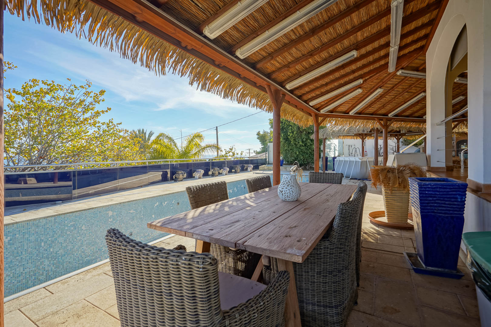 Covered terrace featuring a rustic wooden dining table and wicker chairs under a thatched pergola, situated beside the mosaic-tiled swimming pool.