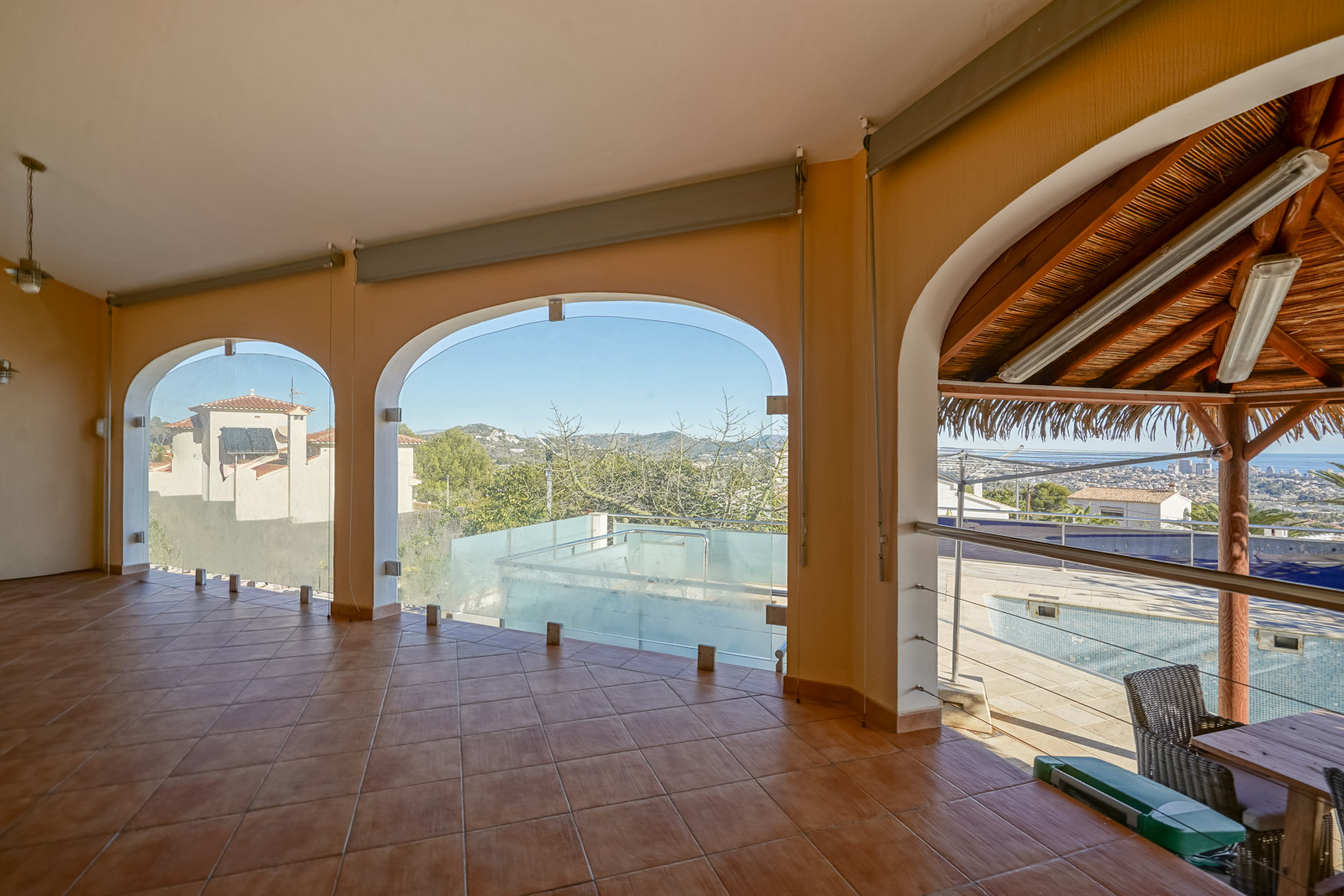 Covered terrace featuring terracotta tile flooring and arched openings with glass enclosures overlooking the pool area and Calpe coastline.
